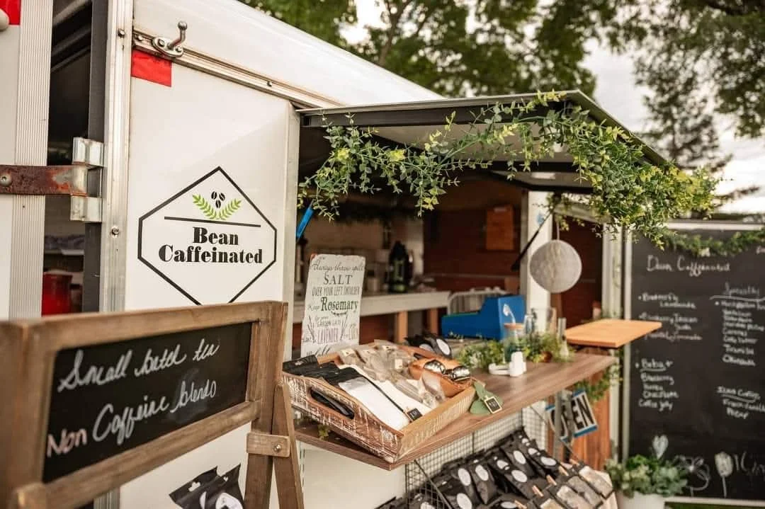 A coffee stand with a sign reading 'Bean Caffeinated' and a chalkboard sign indicating 'Small brew two Non Caffeinated blend.' The stand has a wooden countertop with various coffee-related items, some greenery, and background trees.