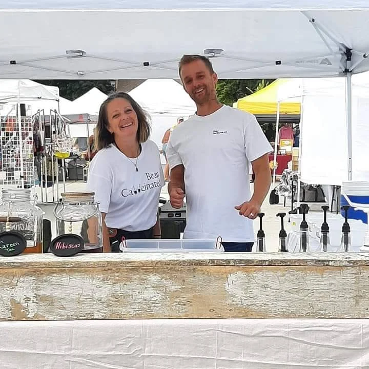 Two smiling people behind a food stall at an outdoor market, wearing white shirts, with jars and bottles on the counter.