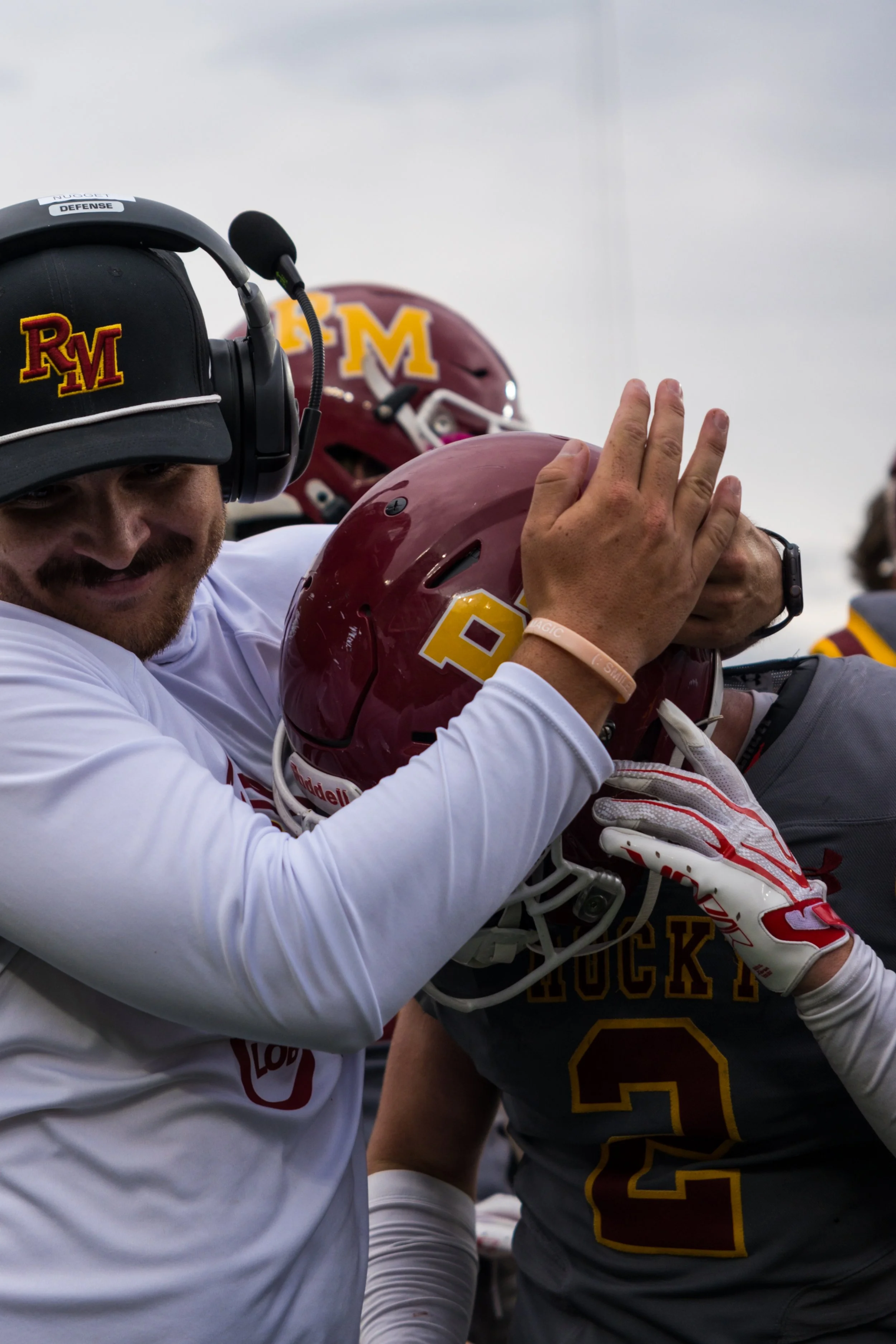 A football coach adjusting a player's helmet during a game, with other team members in the background. The coach is wearing a headset and a black cap with an
