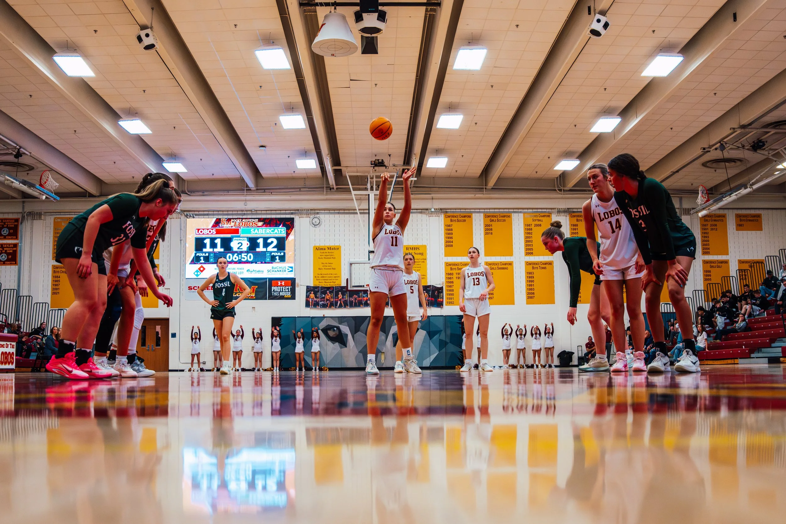 A women's volleyball team practicing or warming up in a gymnasium, with players stretching or preparing on the court. The scoreboard shows 11-12 in the second set with 6 minutes and 50 seconds remaining. The gym has yellow banners on the wall and a large digital scoreboard.