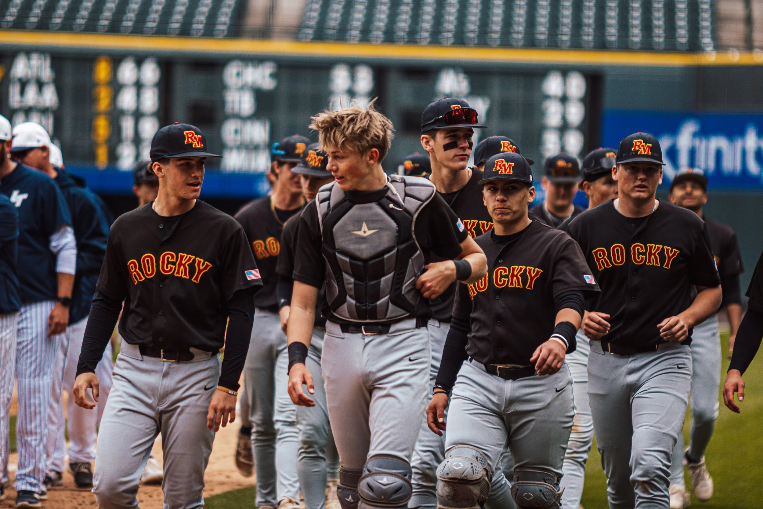 A group of baseball players walking on the field, dressed in black and gray uniforms with "Rocky" written on their shirts, some wearing caps with the initials "RM," and smudged eye black.