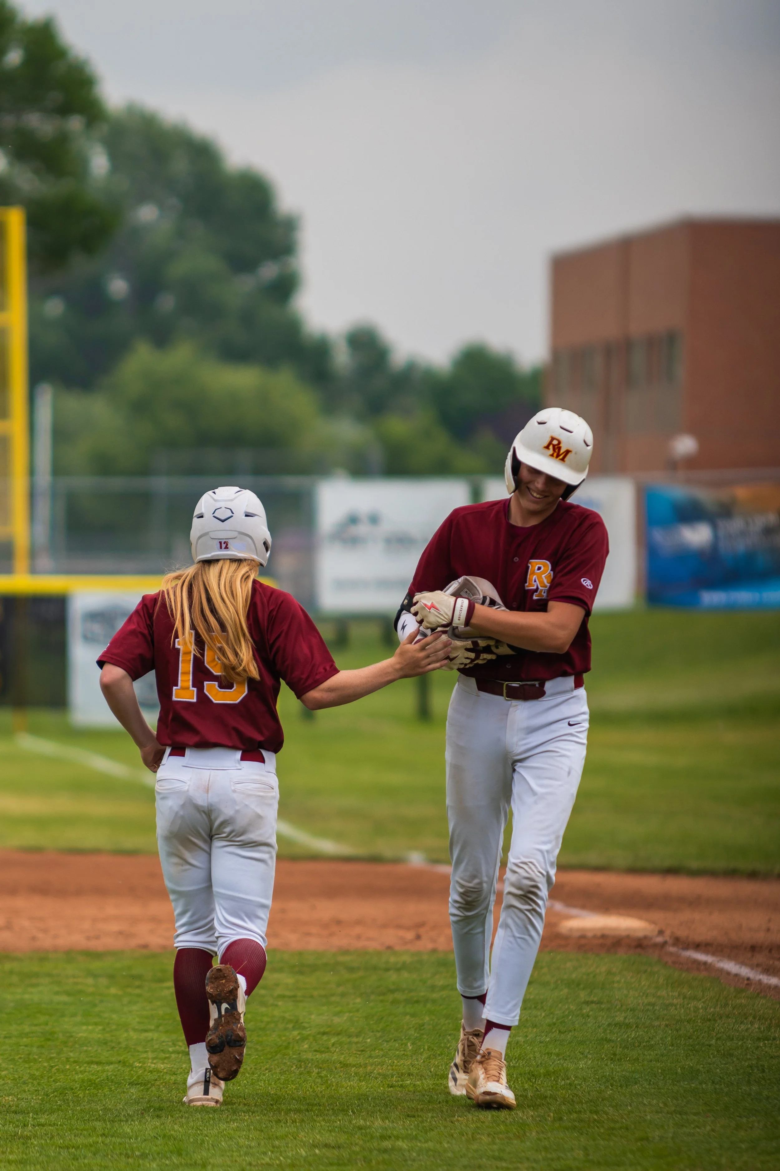 Two young female softball players in maroon jerseys and white pants walking on the field. The girl on the right is smiling and holding a glove, while the girl on the left has her back to the camera. They are wearing white helmets, and the girl on the left has the number 15 on her jersey.
