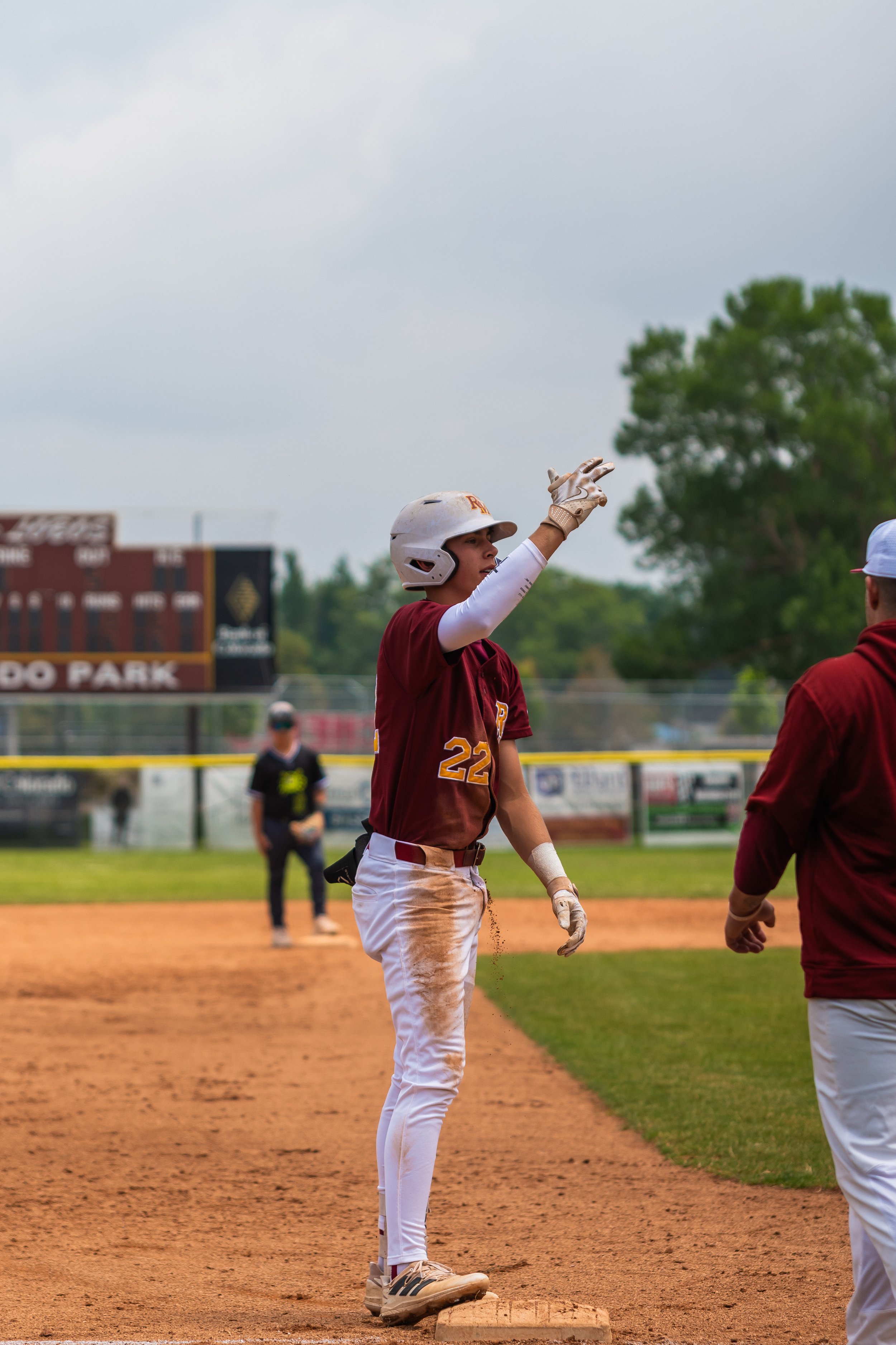 A young baseball player in a maroon uniform and white helmet points towards the field while standing on a base, with other players and a scoreboard in the background.