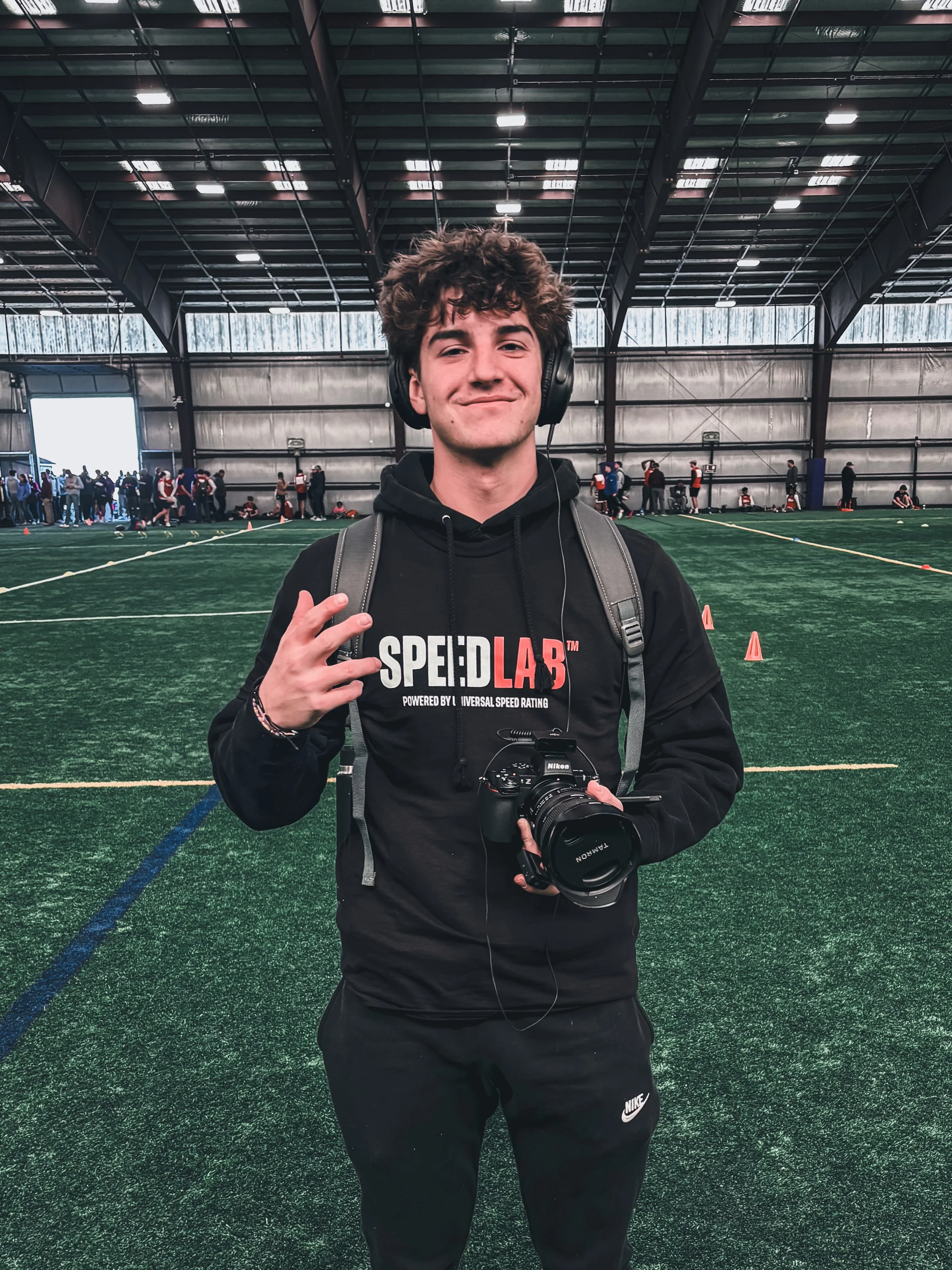 A young man with curly hair smiling at the camera inside an indoor sports facility, wearing a black hoodie with 'SPEEEDLAB' logo, headphones, and holding a camera, with a group of people and sports activities in the background.