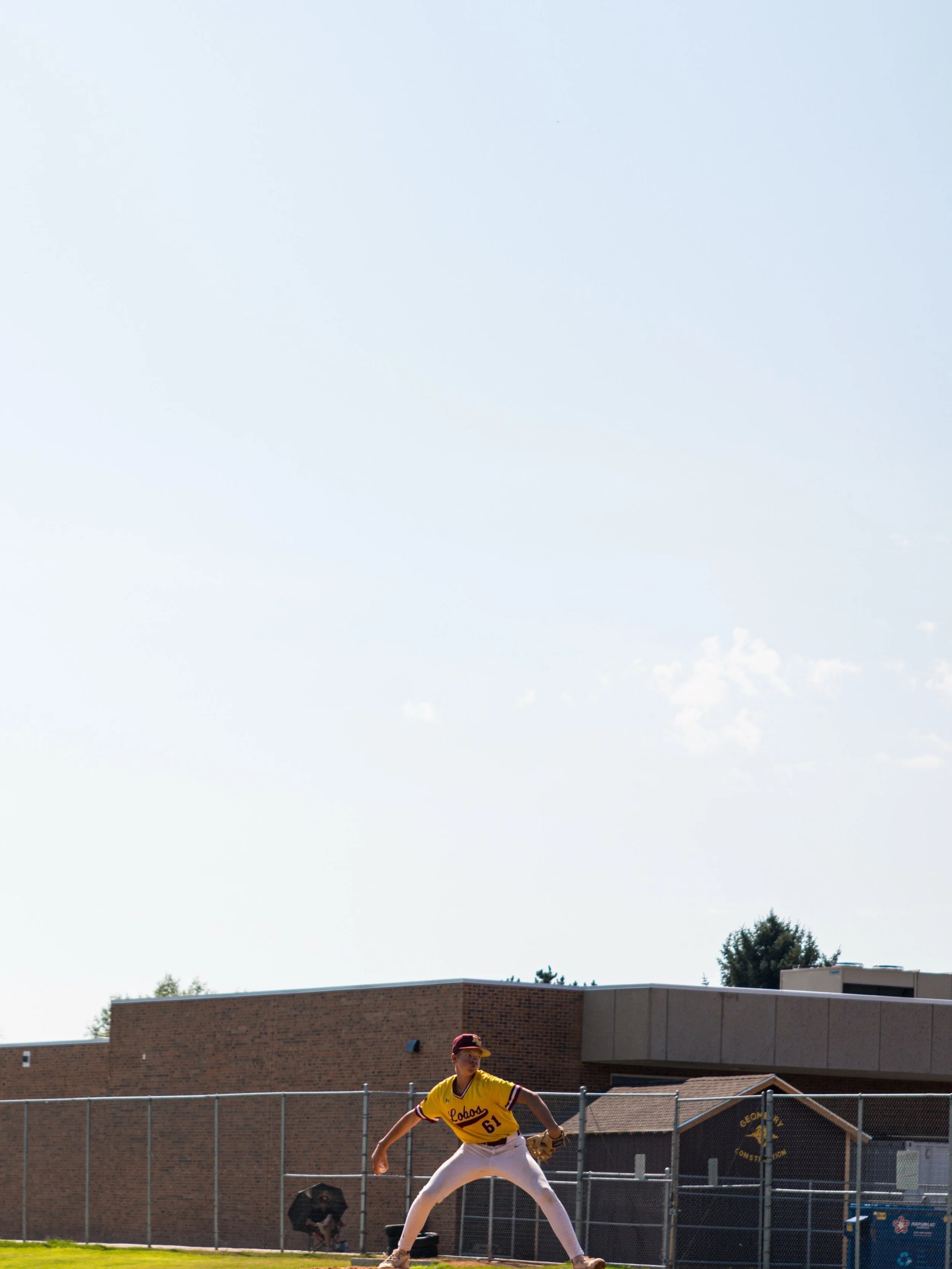 A baseball player wearing a yellow and maroon uniform is in a ready position on the field. He is wearing a maroon cap and has a glove on his left hand. He is on a baseball field with a chain-link fence and a school building in the background, with trees and a clear sky.