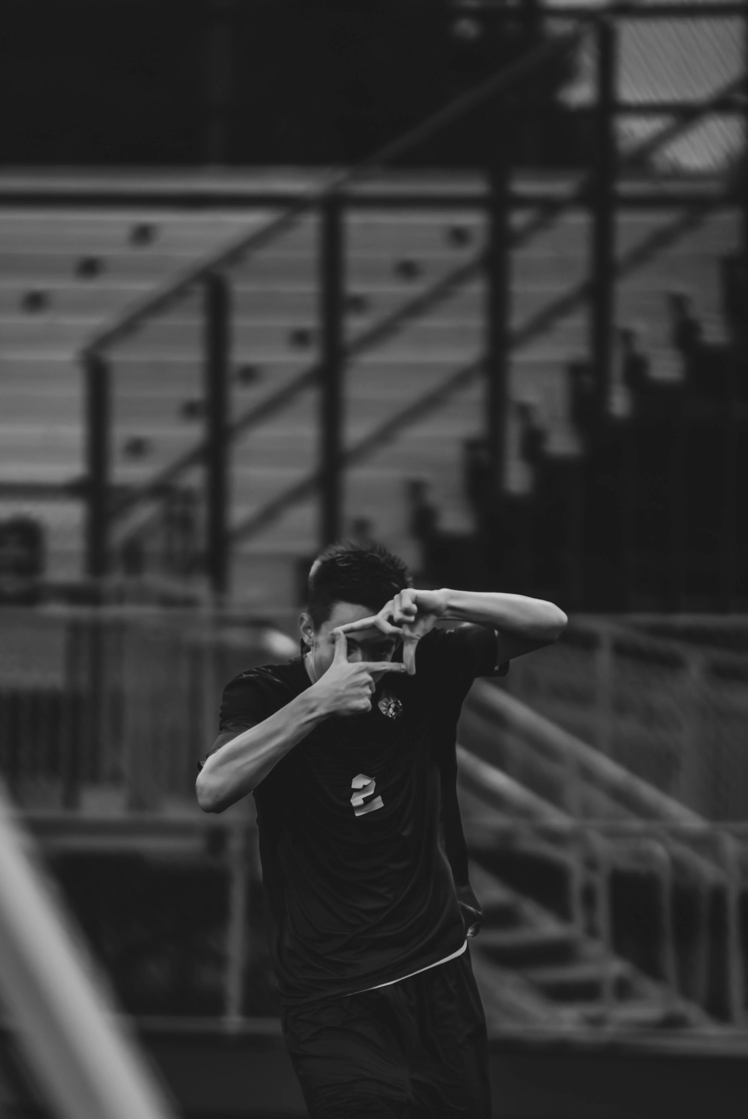 A young male athlete wearing a sports jersey, making a heart shape with his hands in front of a sports facility with a metal staircase in the background.