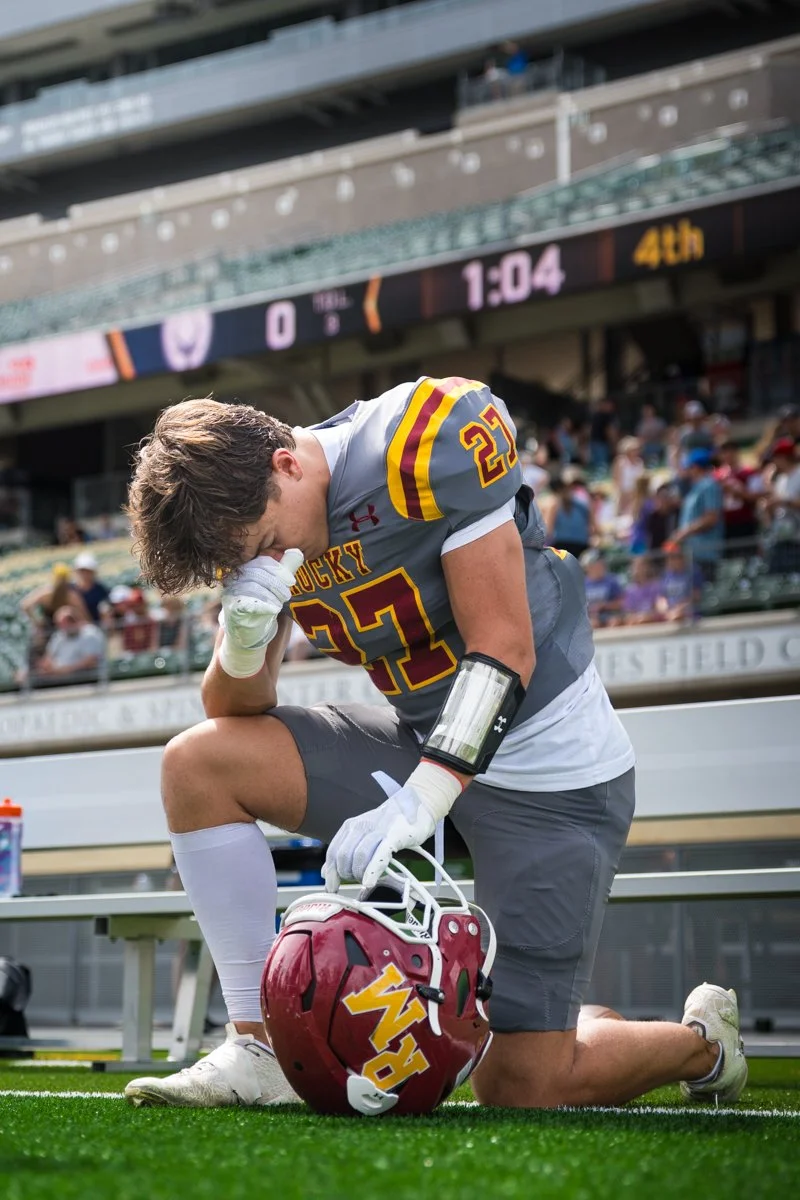 A football player kneeling on one knee with head bowed and hand on his forehead in the stadium, holding a red helmet with a yellow logo, during a game with a digital scoreboard showing zero points and 1 minute 4 seconds left in the fourth quarter.