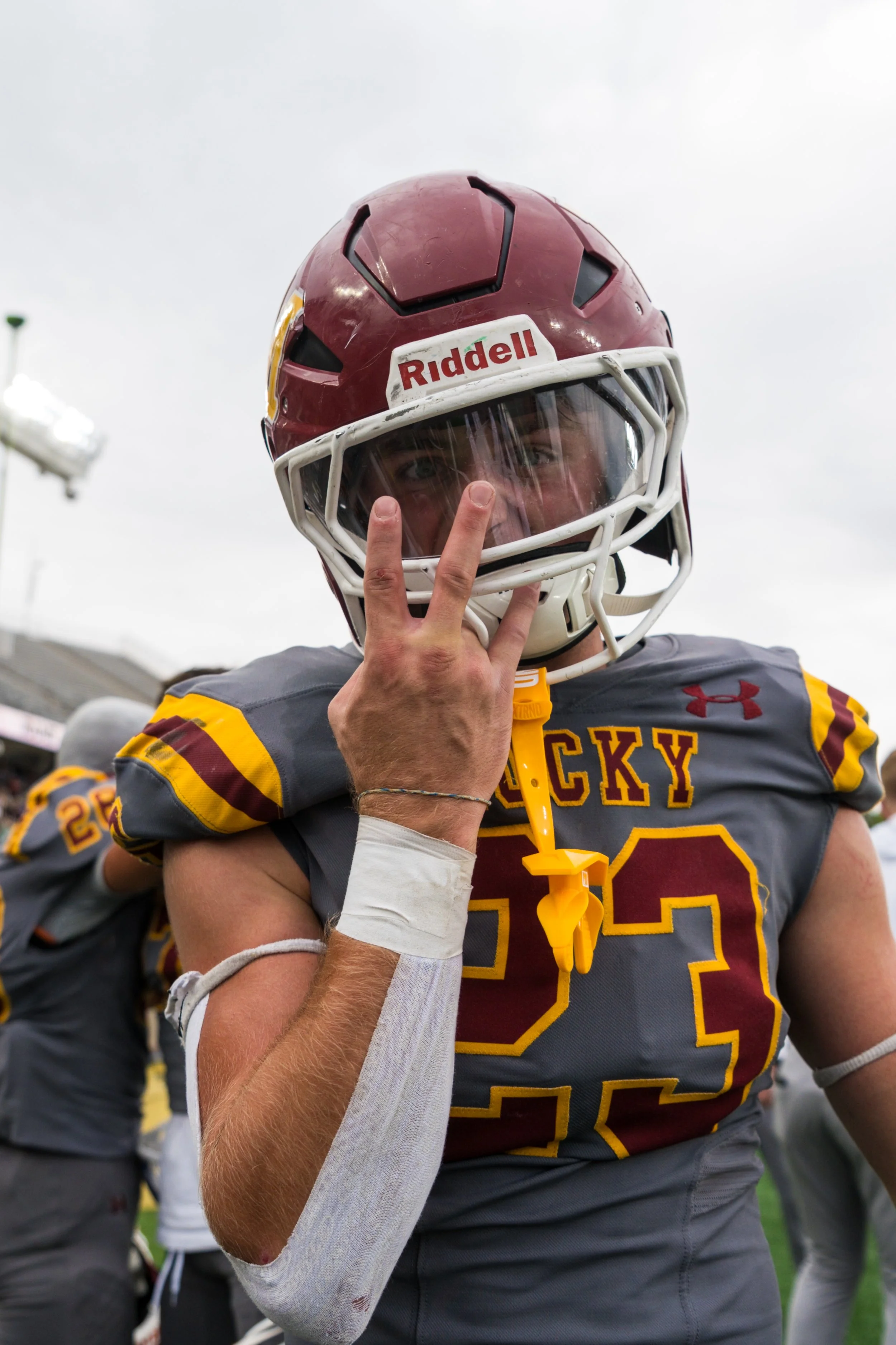 A football player in a gray and maroon uniform with the number 32, wearing a maroon helmet with a white faceguard, making a peace sign with his fingers
