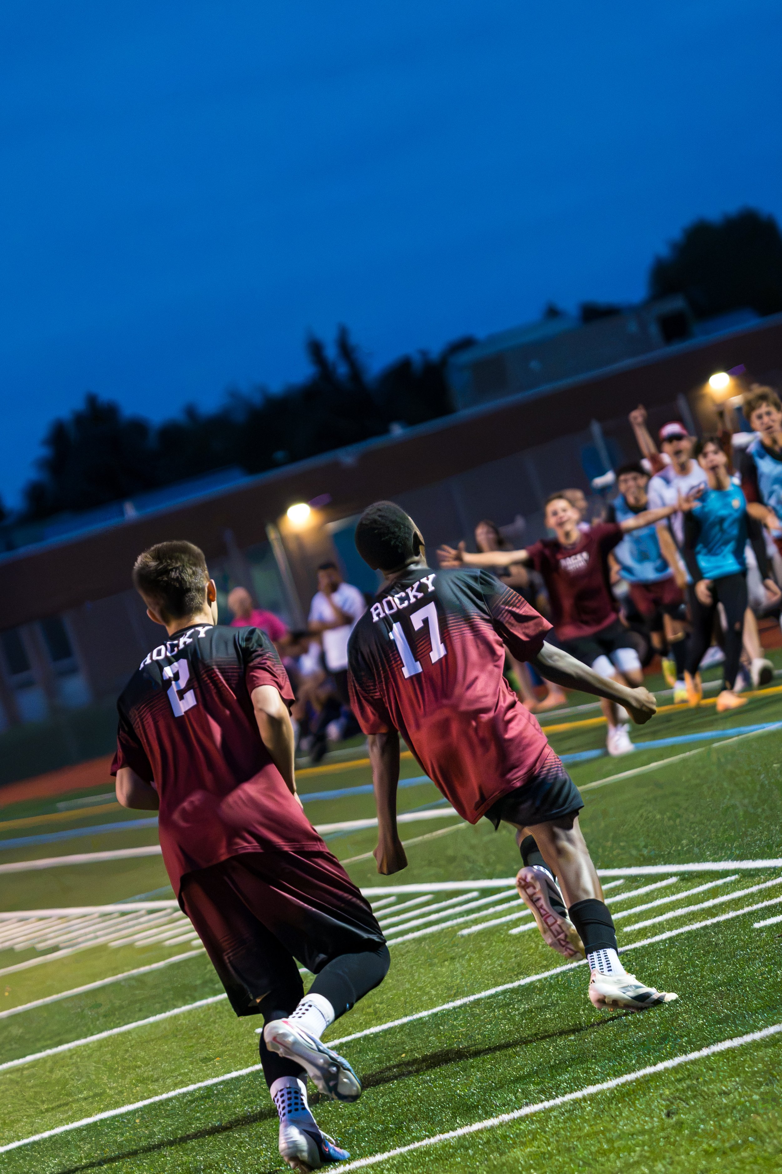 Young boys playing soccer on a field during the evening with a crowd of spectators watching in the background.
