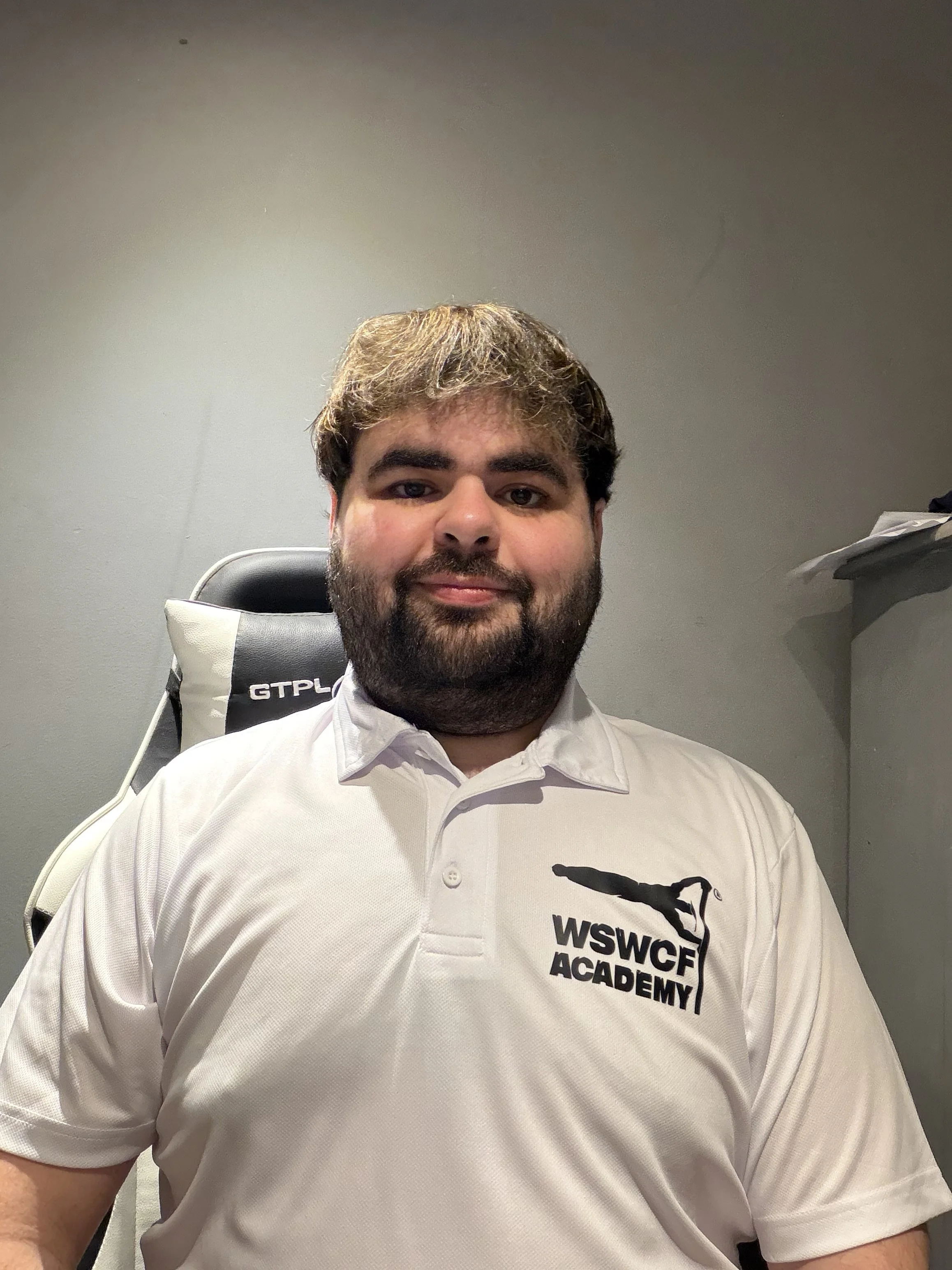 A man with brown hair and a beard is sitting in a chair, wearing a white polo shirt with the WSWCF Academy logo. The background is a plain gray wall.