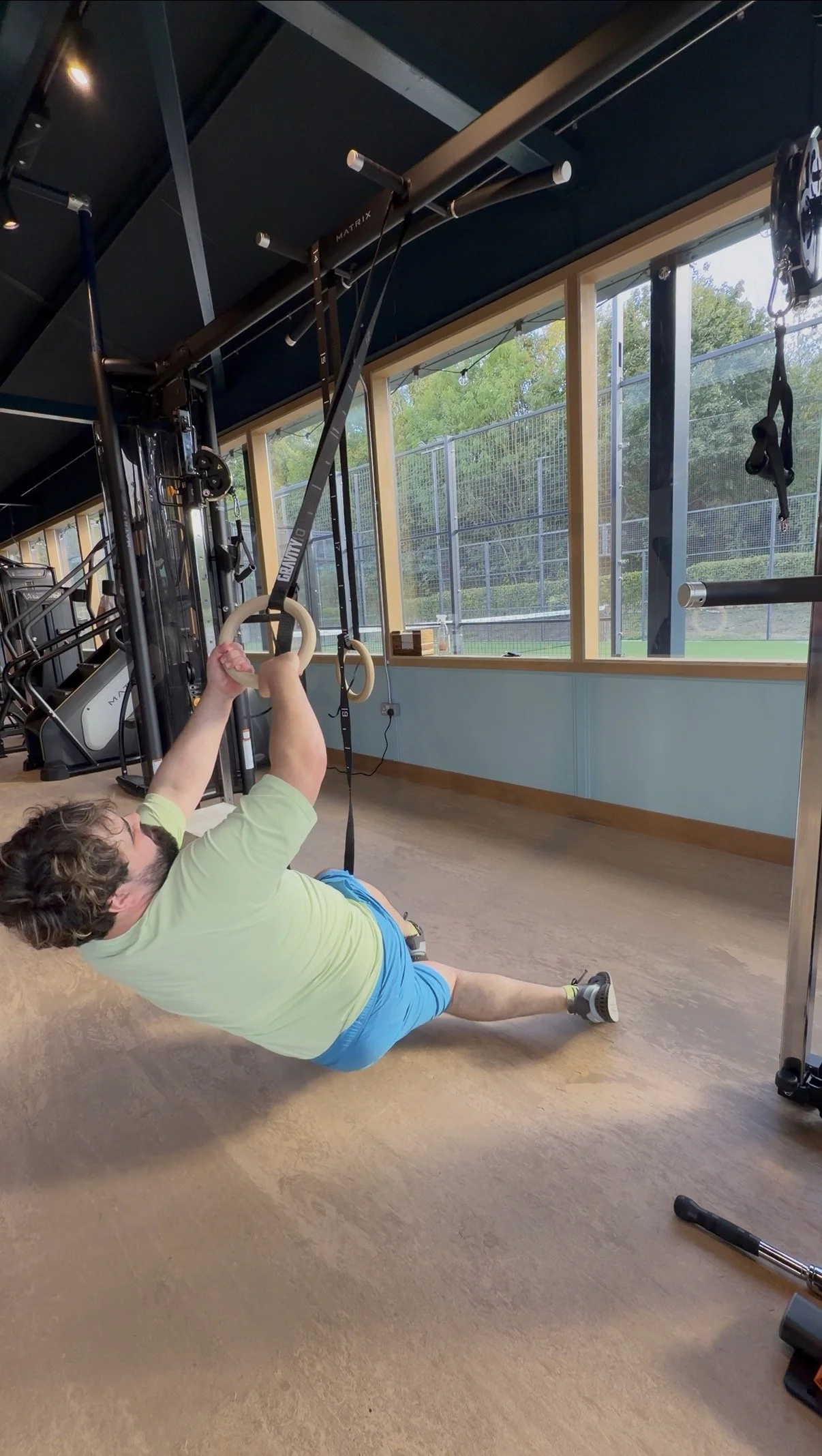 Man performing an exercise on gym floor, holding gymnastic rings attached to a ceiling-mounted structure inside a gym with large windows showing outdoor greenery.