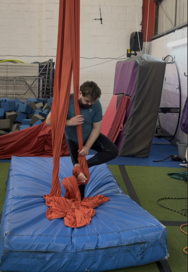 A person practicing aerial silks in an indoor gym, holding onto orange fabric suspended from the ceiling, with a blue crash mat below.