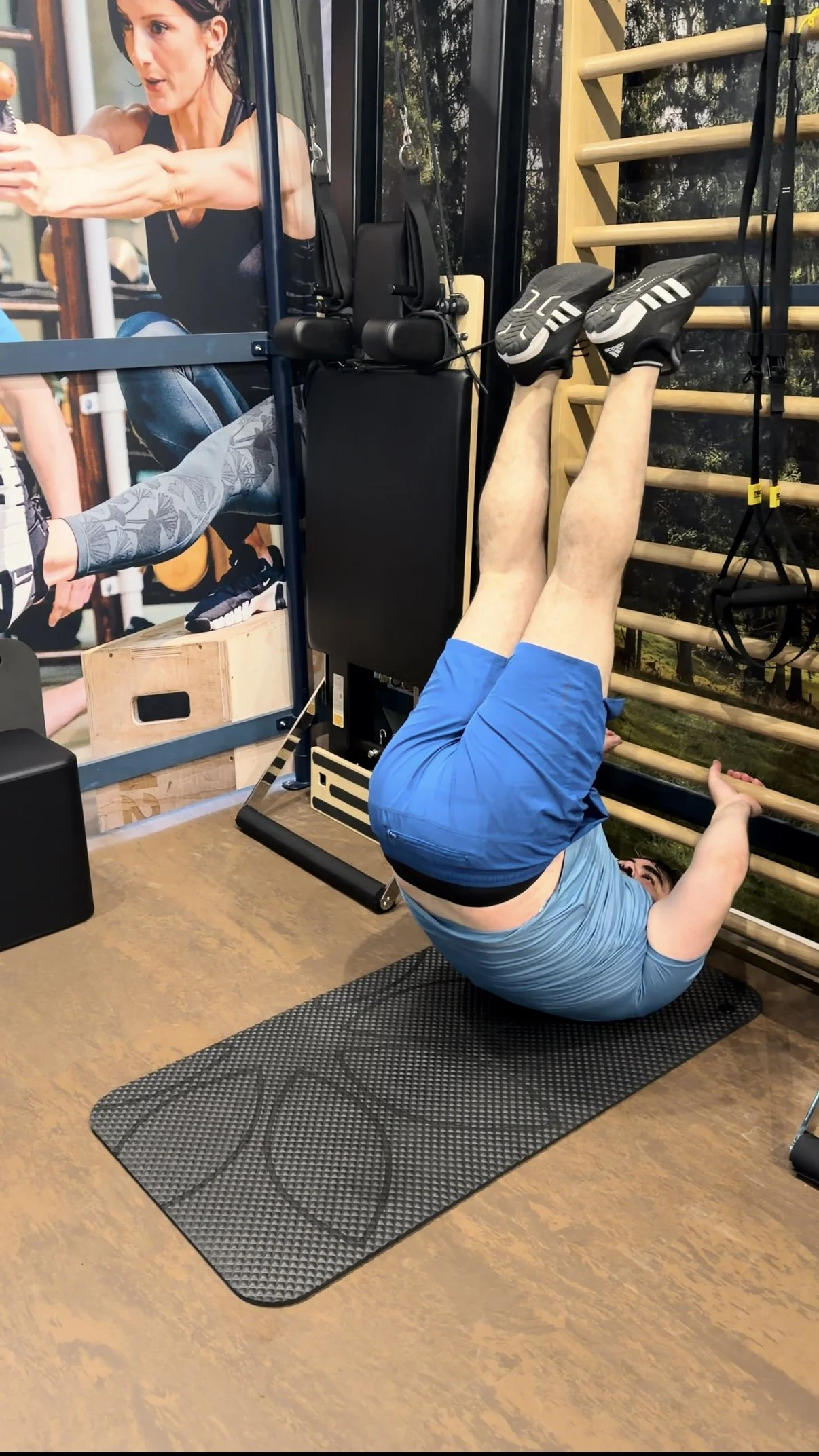 Person in blue shorts and a light blue shirt doing a yoga pose on a black rubber mat in a gym, with gym equipment and a wall mural in the background.
