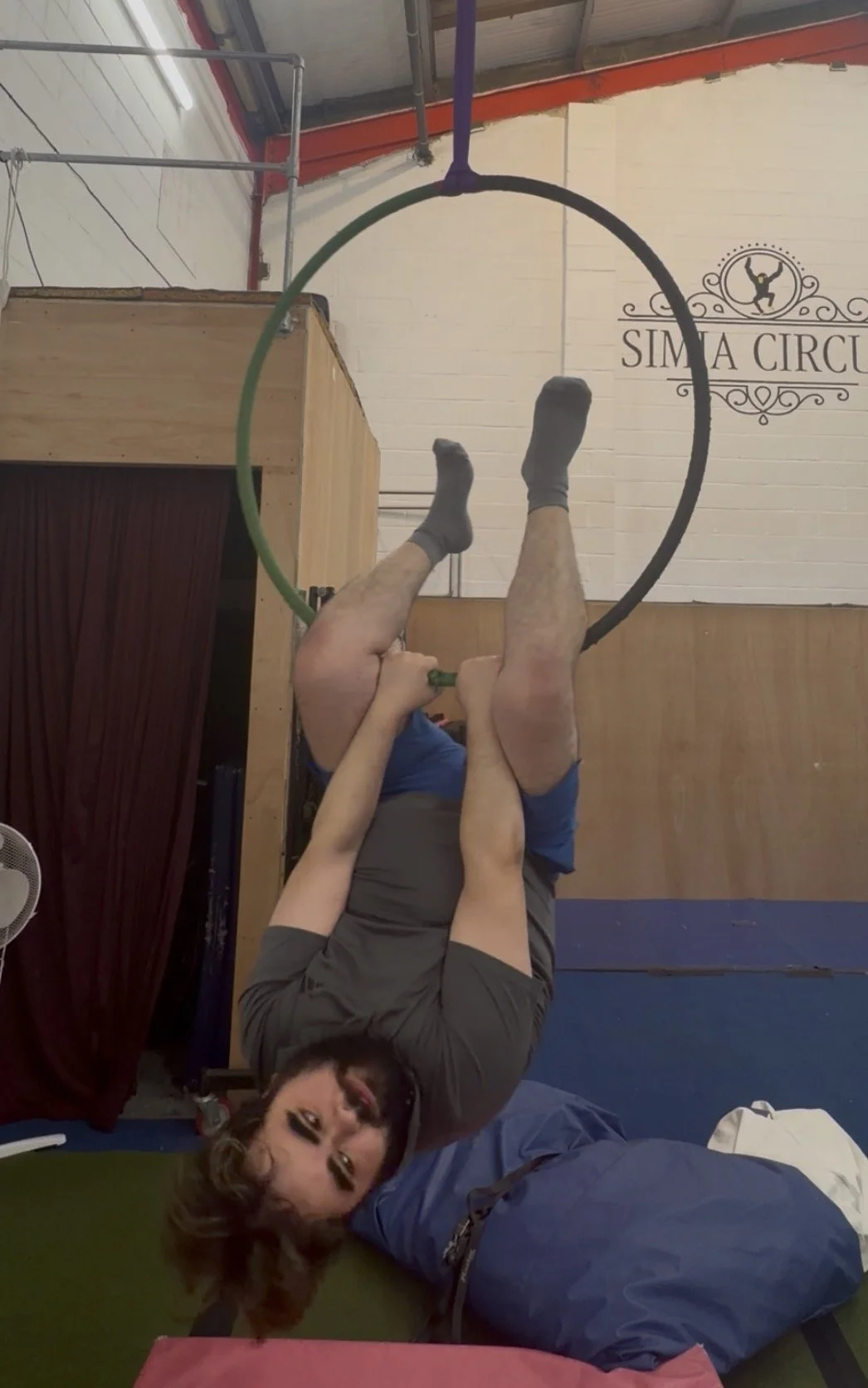 A woman hanging upside down from a gymnastic ring, holding onto a child's arm. The woman has makeup on and is smiling at the camera. The background shows a wall with a logo that reads 'SIMA CIRCUS' and an indoor gym setting.