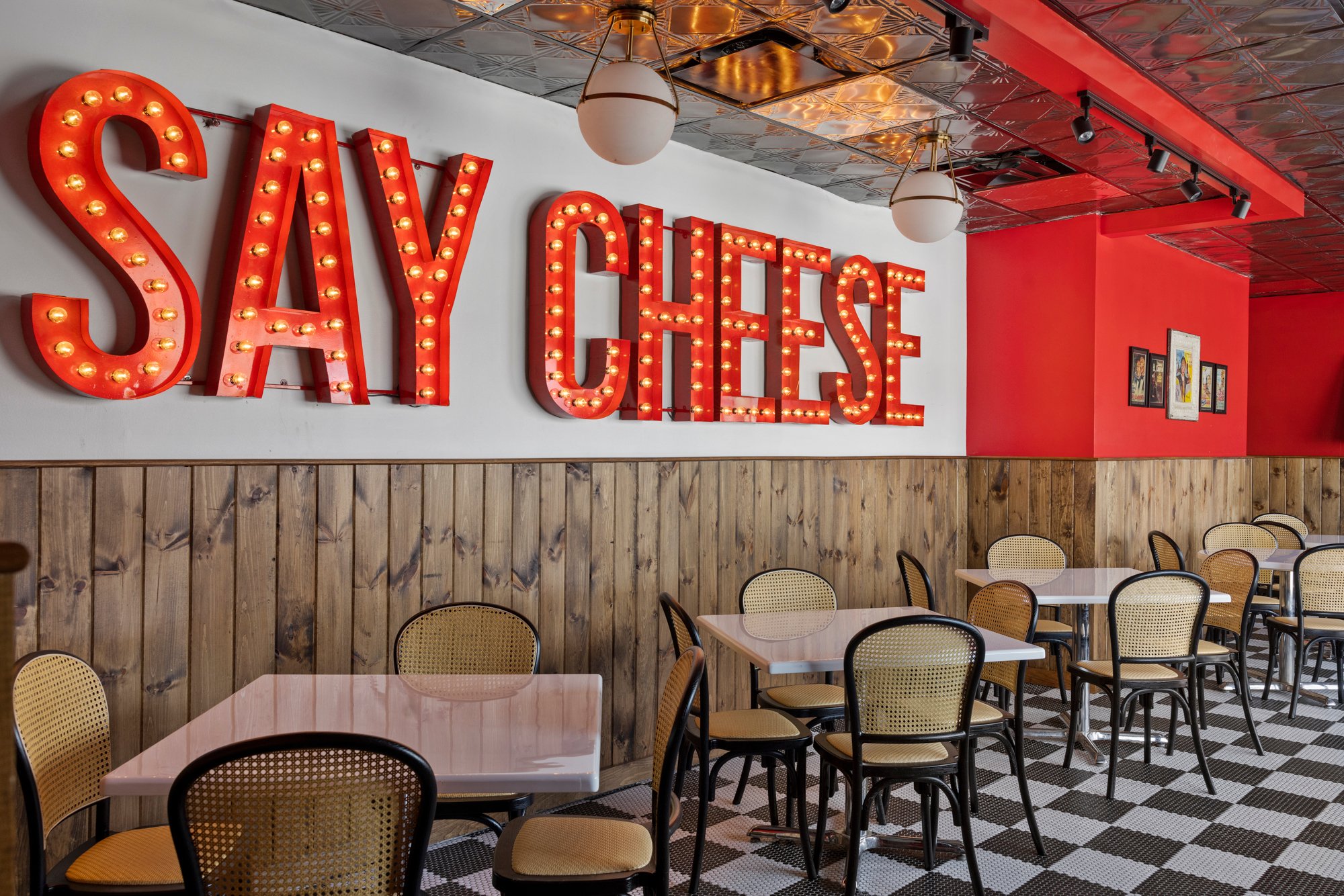 Interior of a restaurant with a bright red and white color scheme, featuring large illuminated 'SAY CHEESE' sign on the wall, wooden paneling, round ceiling lights, patterned ceiling tiles, framed pictures, and tables with chairs.