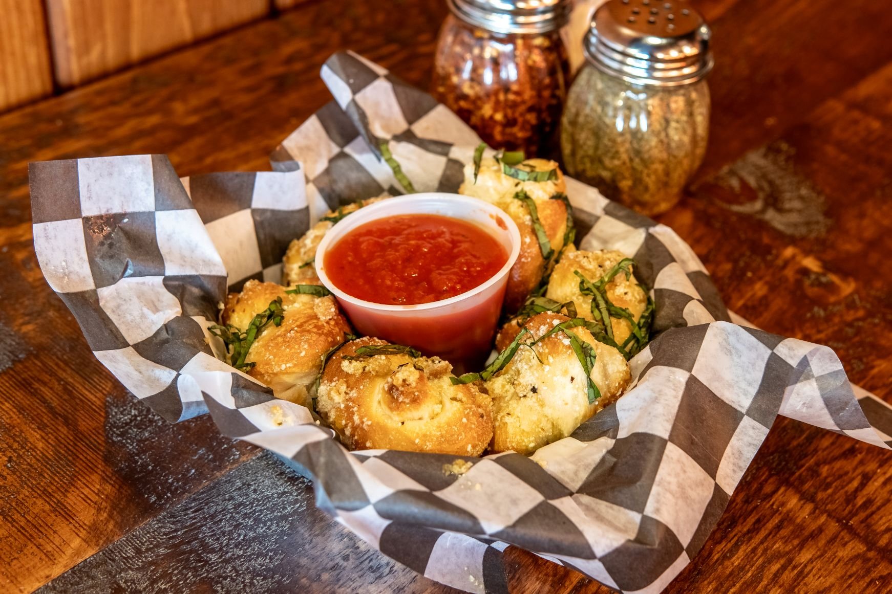 Basket of fried cheese balls garnished with chopped green herbs, served with a cup of red marinara sauce, on checkered paper in a restaurant. Salt and pepper shakers are in the background.