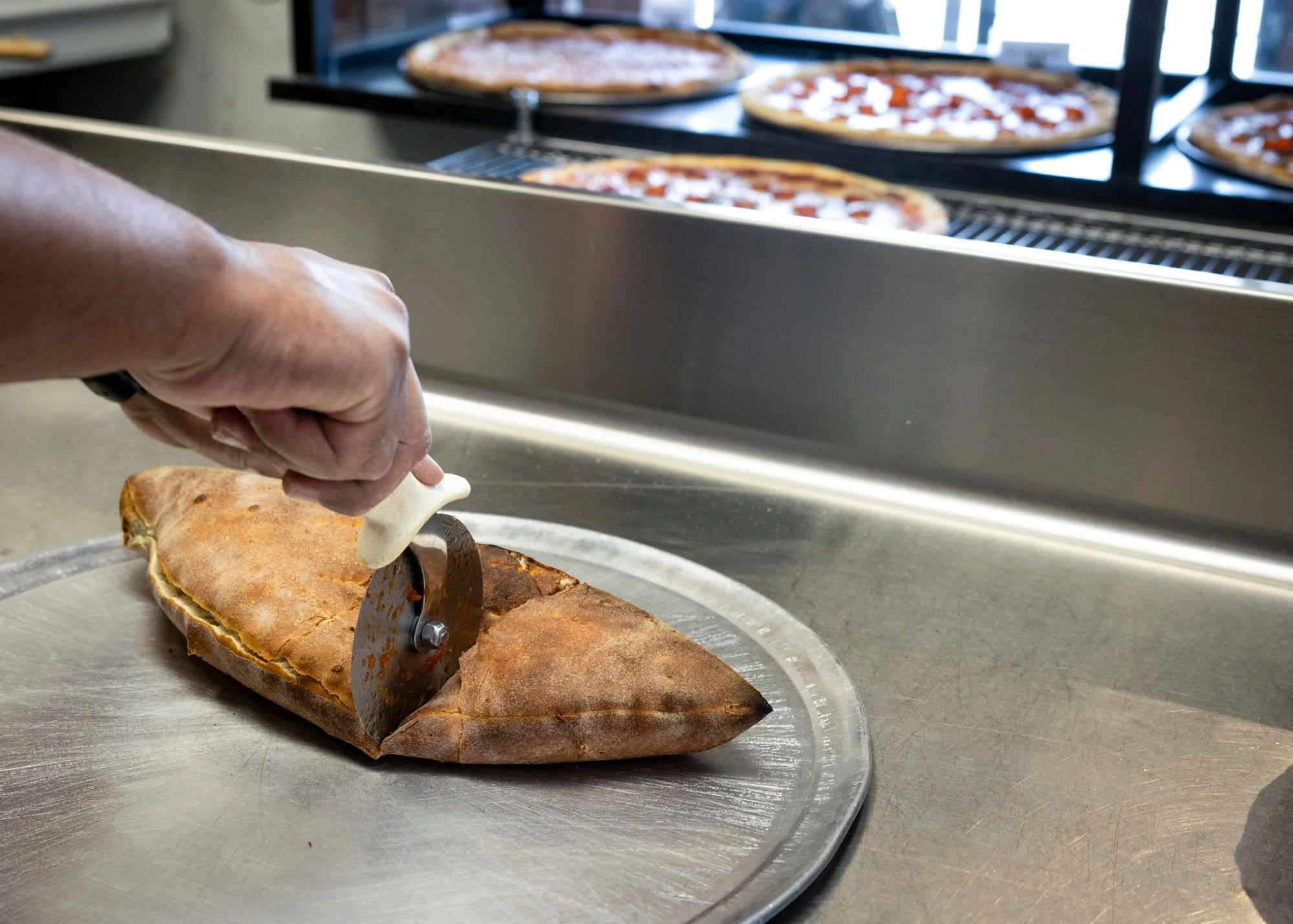 A person slicing a large cooked fish with a round slicing tool on a metal tray, with multiple pizzas baking in the oven in the background.