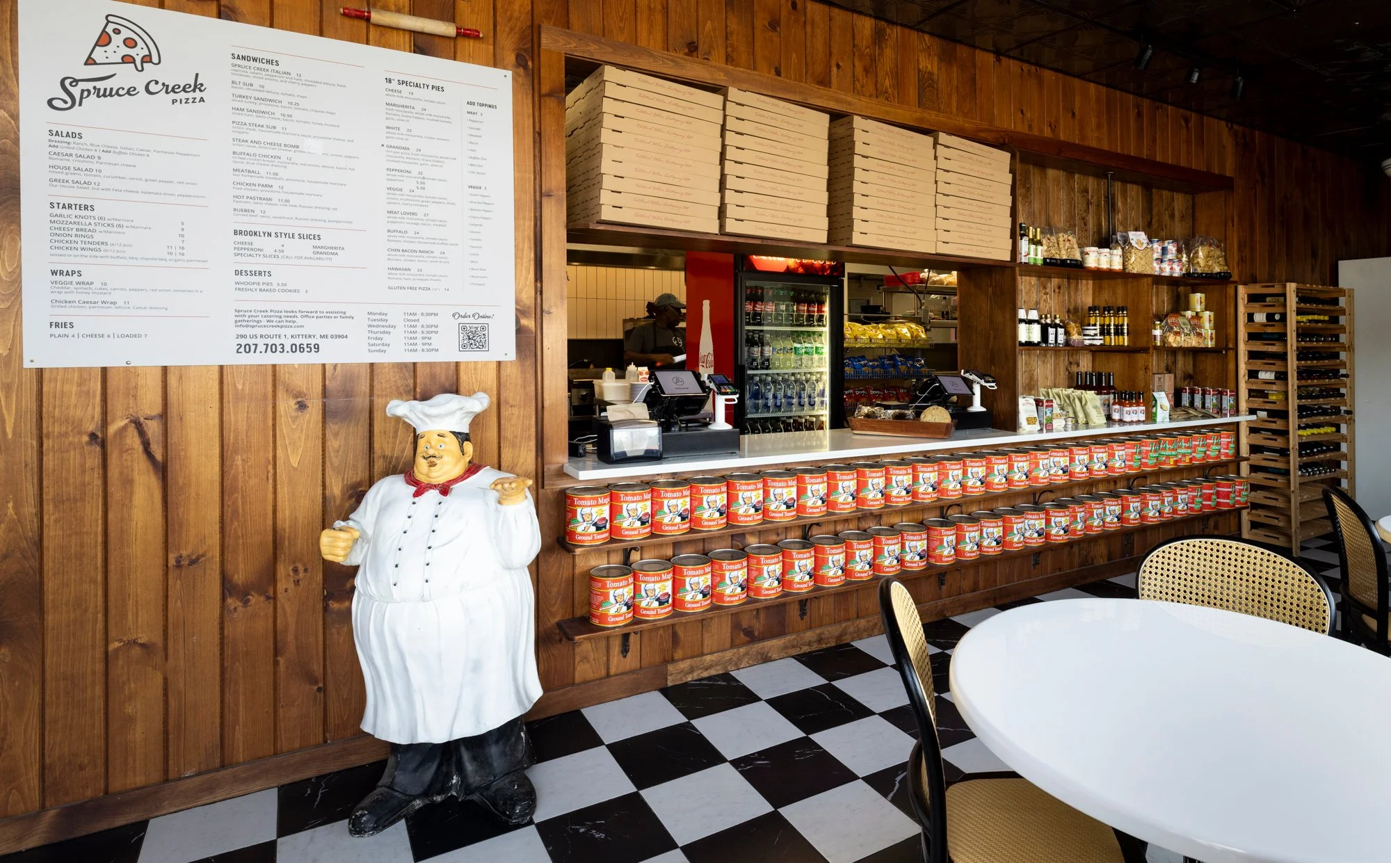 Interior of a restaurant with a snack counter, featuring a large menu board, stacked pizza boxes, canned tomatoes, and various condiments on shelves. A cheese chef statue stands next to the counter, and tables with chairs are in the foreground.