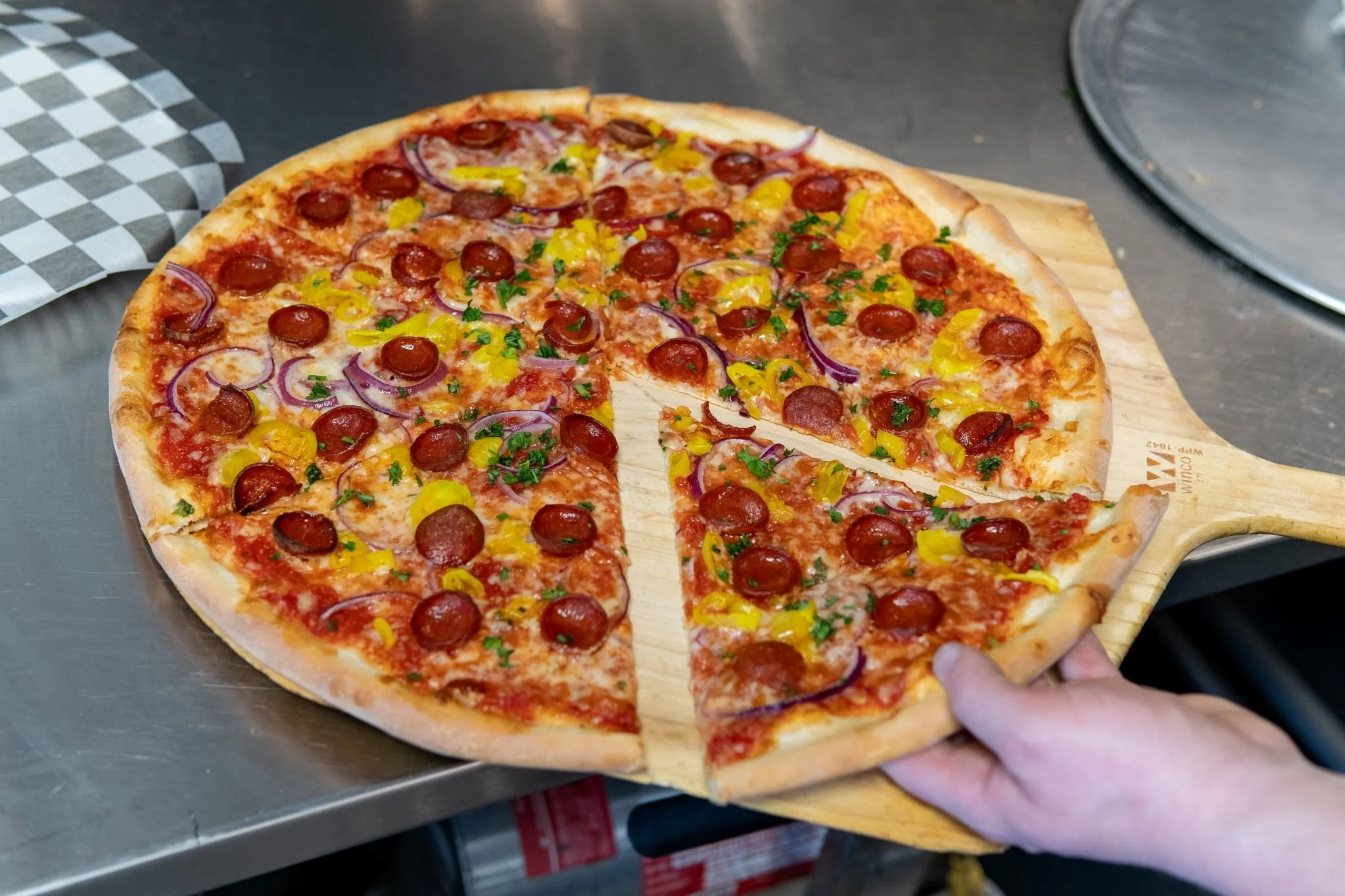A hand holding a slice of pepperoni pizza with a variety of toppings, including red onions, yellow peppers, and green herbs, served on a wooden pizza paddle.