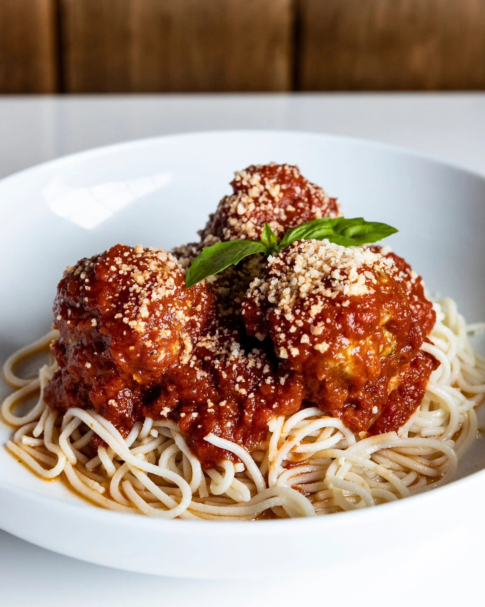 Spaghetti with meatballs topped with tomato sauce, grated cheese, and a basil leaf in a white bowl.