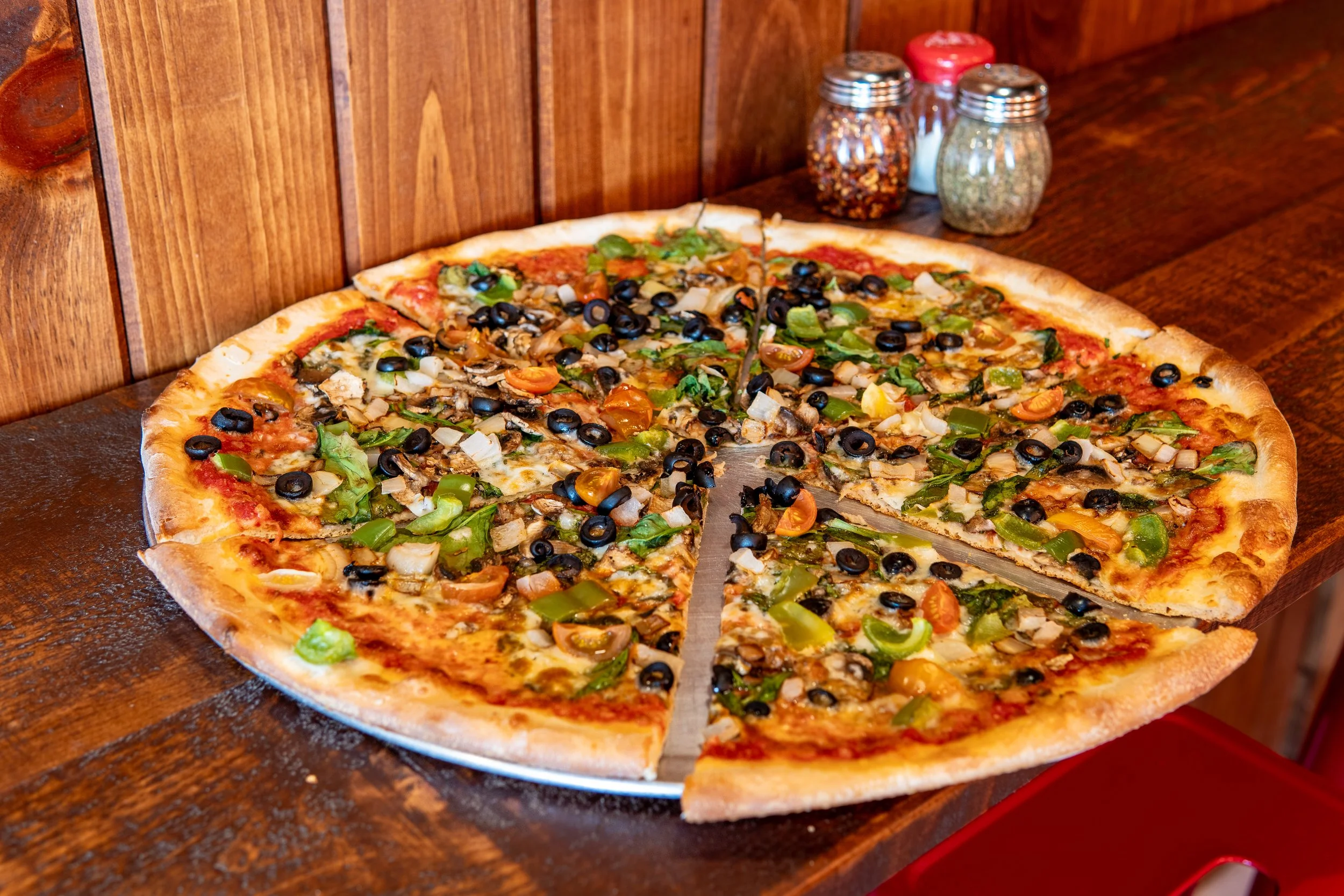 A large pizza with toppings including black olives, green bell peppers, onions, and cherry tomatoes on a wooden table in a restaurant setting.