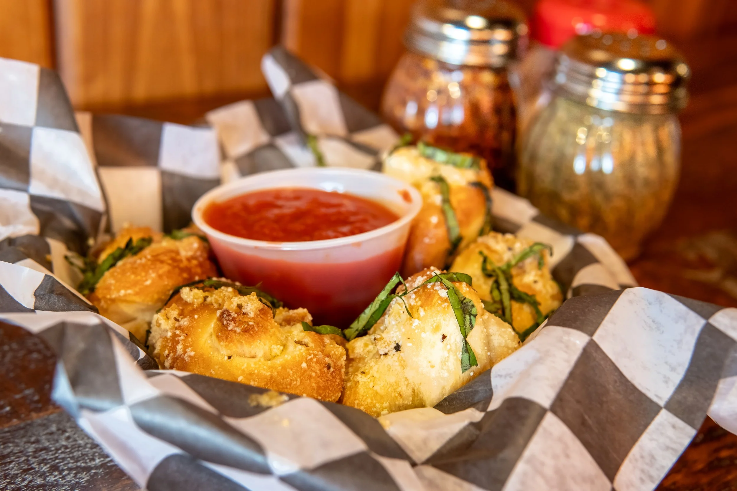 Fried mozzarella sticks with basil garnish, marinara sauce in a small cup, and two jars of spices in the background on a dark surface.