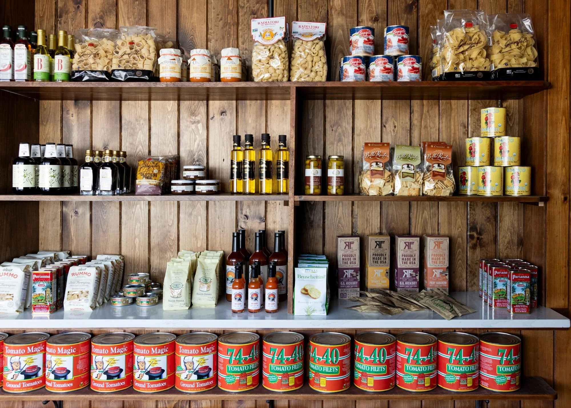 Wooden shelves stocked with various canned and packaged food products, including pasta, tomato sauce, olive oil bottles, and canned tomatoes.