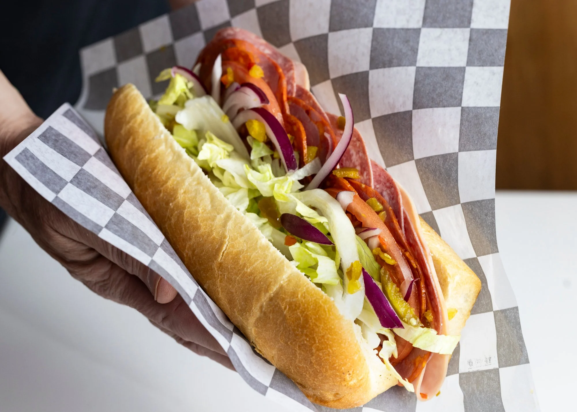 A hot dog with lettuce, tomato, onions, pickles, mustard, and chili, served in a bun on checkered paper.