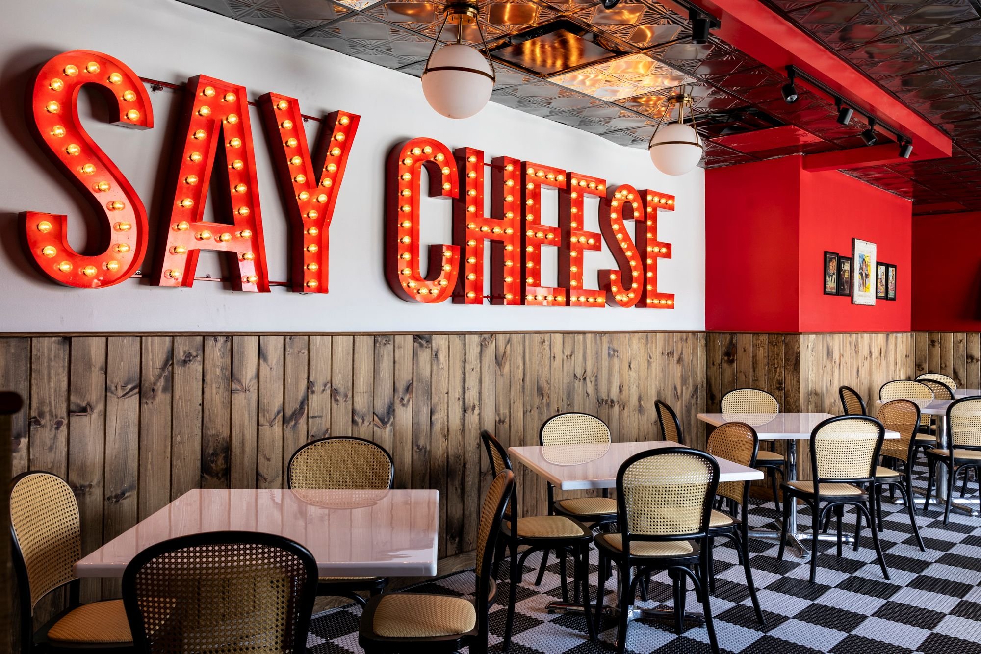 Interior of a restaurant with tables, chairs, a wooden panel wainscoting, vintage style decoration, and a large illuminated sign that reads 'SAY CHEESE' on the wall.