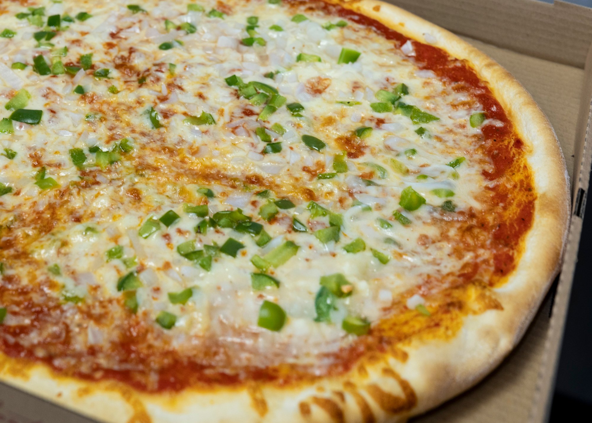 Close-up of a cheese pizza with green peppers and onions on a cardboard box.