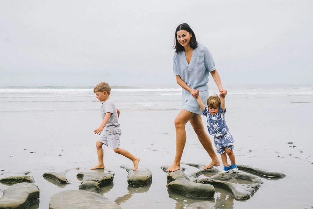 A woman and two children walking on rocks in the ocean on a cloudy day.