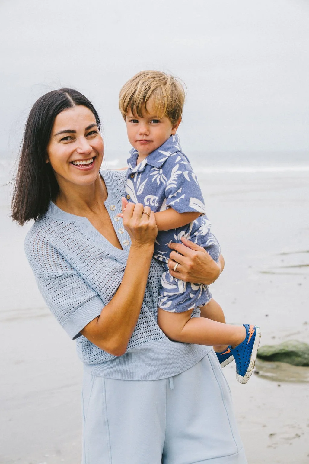 A woman with dark hair and a young boy with light hair smiling at the camera at the beach.
