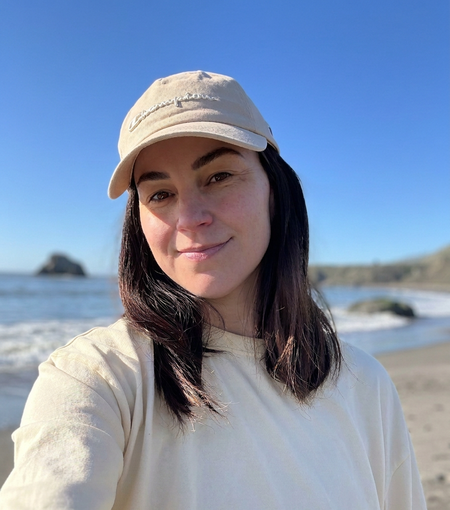 Woman with dark hair wearing a beige cap and cream-colored shirt on a beach with the ocean and rocks in the background on a sunny day.