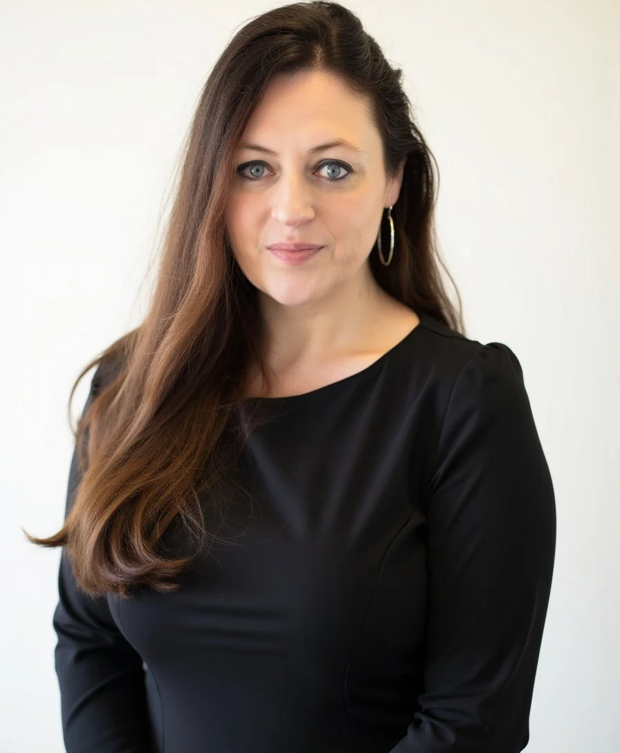 A paralegal with long brown hair and blue eyes, wearing a black top and hoop earrings, looking at the camera with a slight smile, standing against a light background. The paralegal is very experienced with guardianship law.
