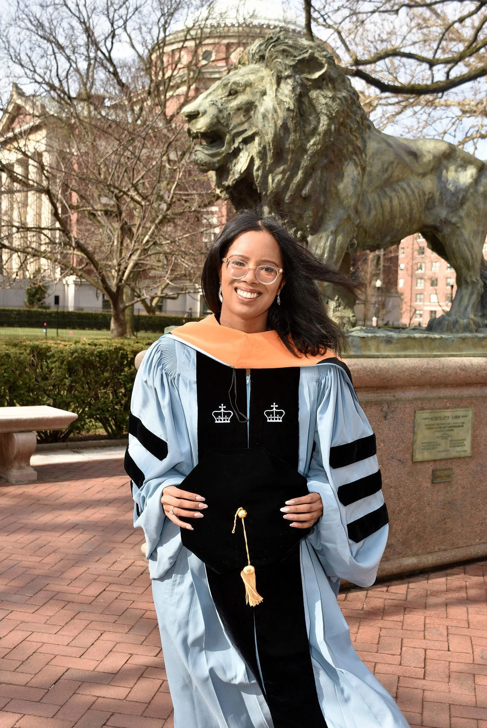 Dr. Justinna Dixon in Columbia University doctoral regalia holding her tam in front of the Columbia Lion statue