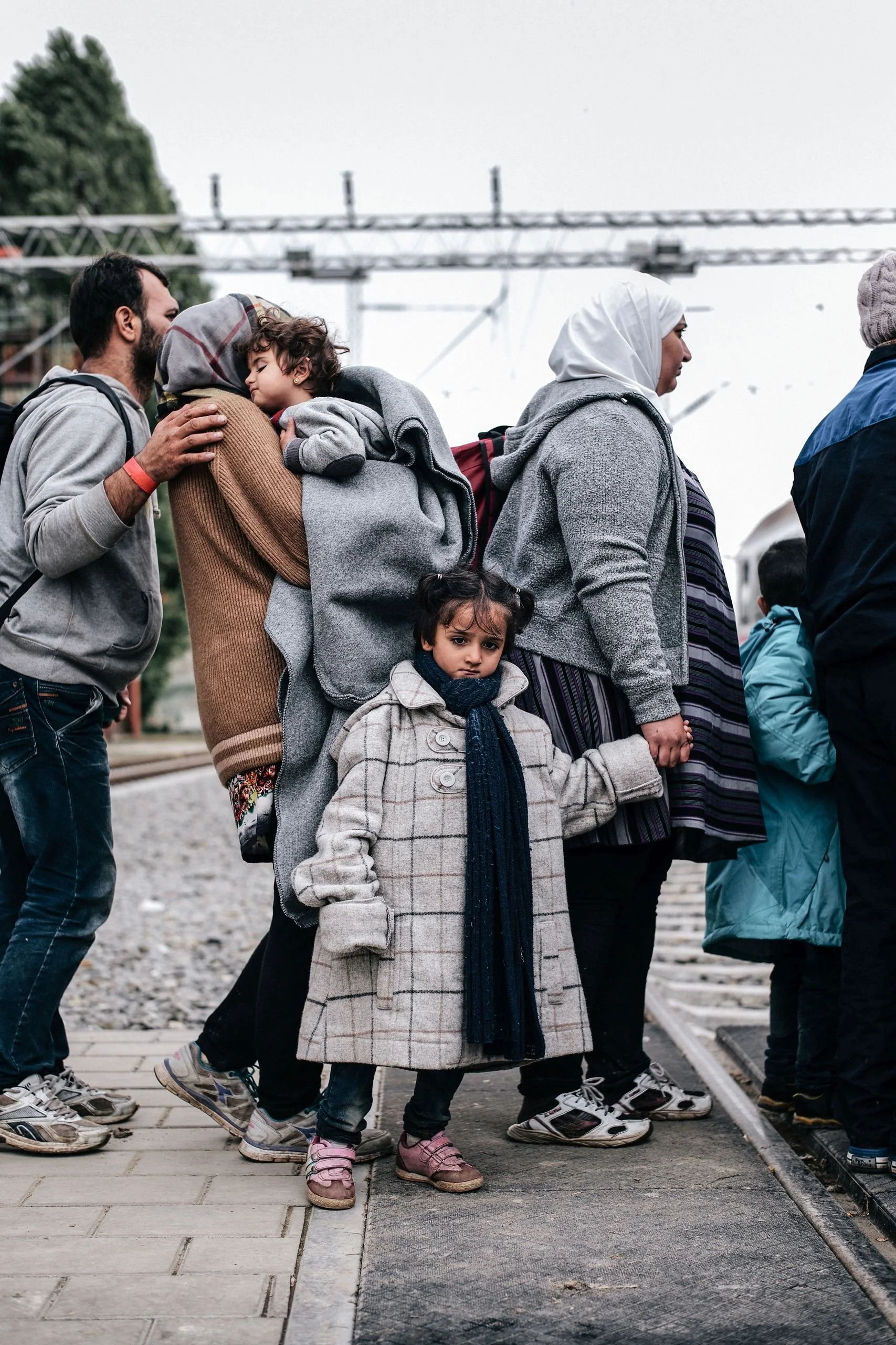 Line of diverse people, including children and adults, crossing a train track outdoors on a cloudy day.