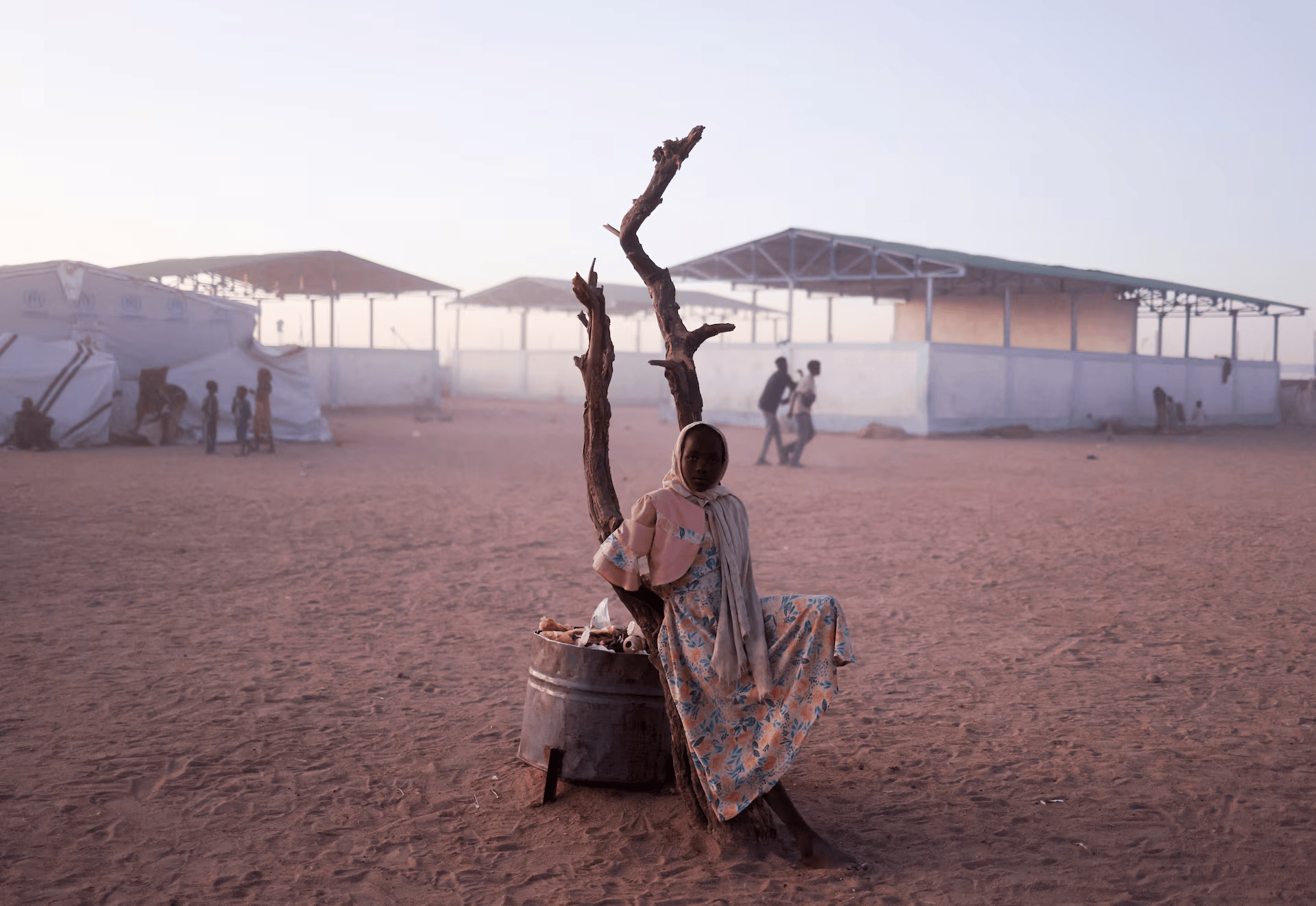 A young girl in a patterned dress and shawl sitting on a metal barrel, holding a stick, with a piece of cloth over her head, in a dusty outdoor setting with tents and people in the background.