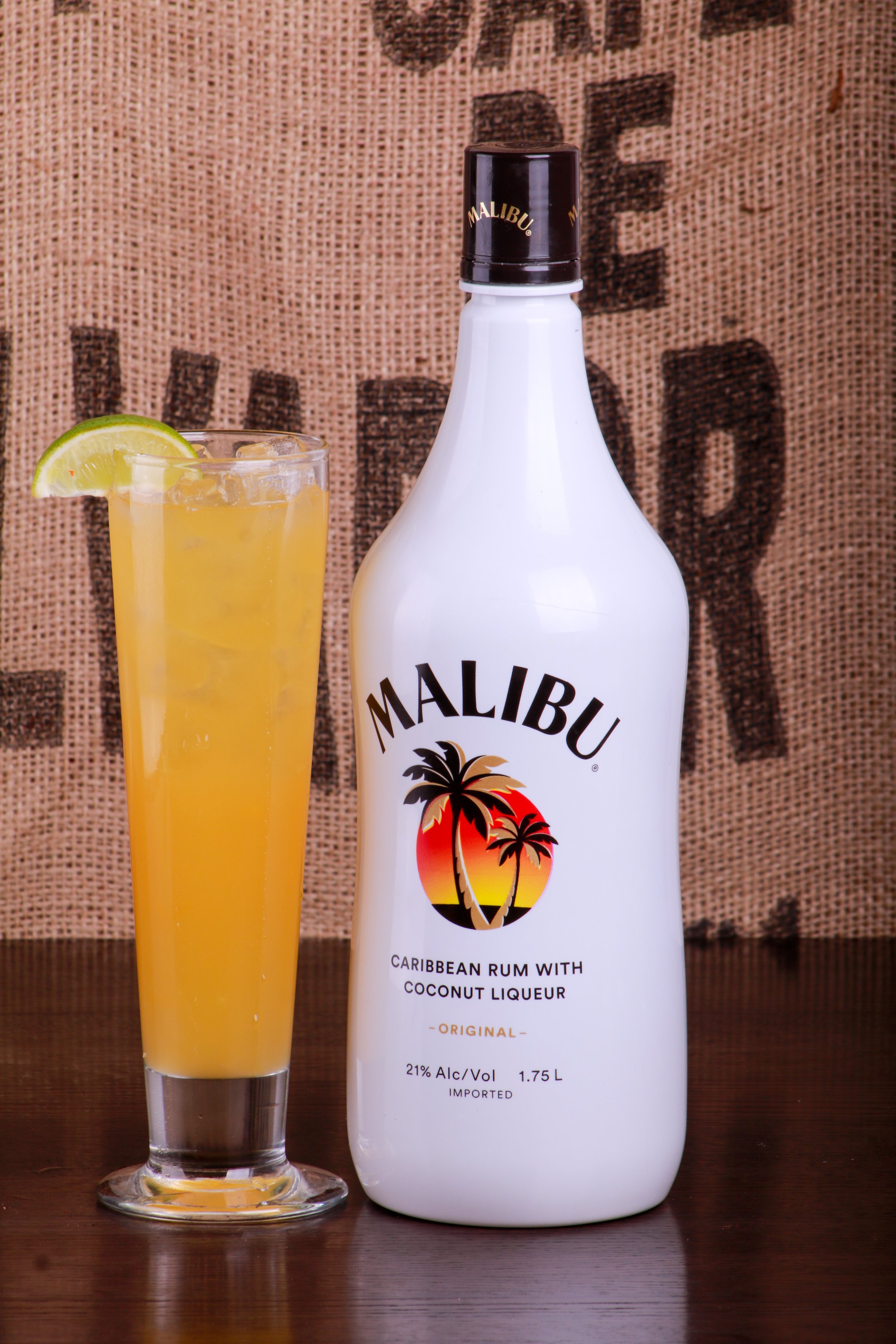 Glass of orange cocktail with lime garnish beside a Malibu coconut rum bottle