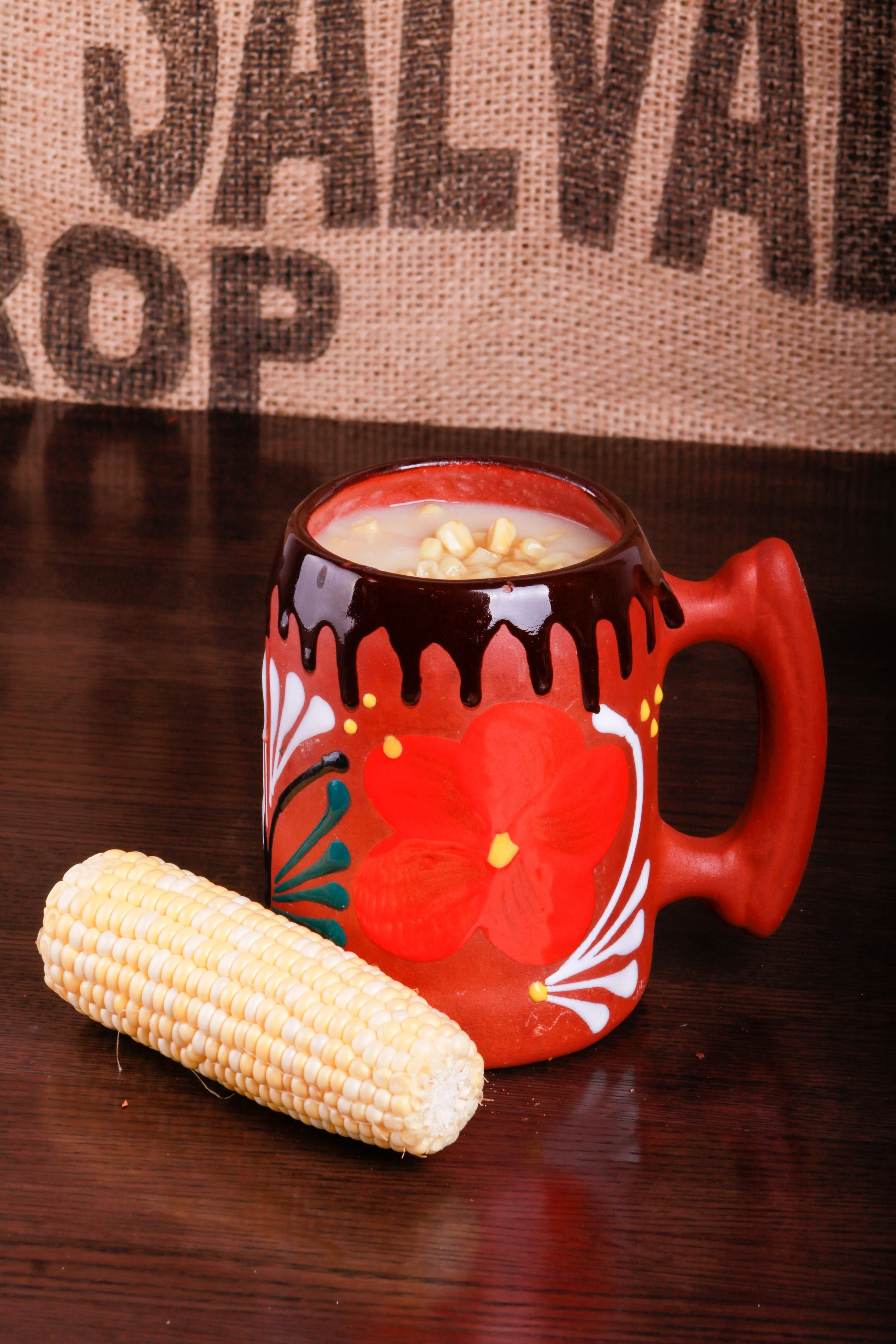 Red decorative mug filled with creamy corn soup, garnished with kernels, beside fresh corn on a wooden table