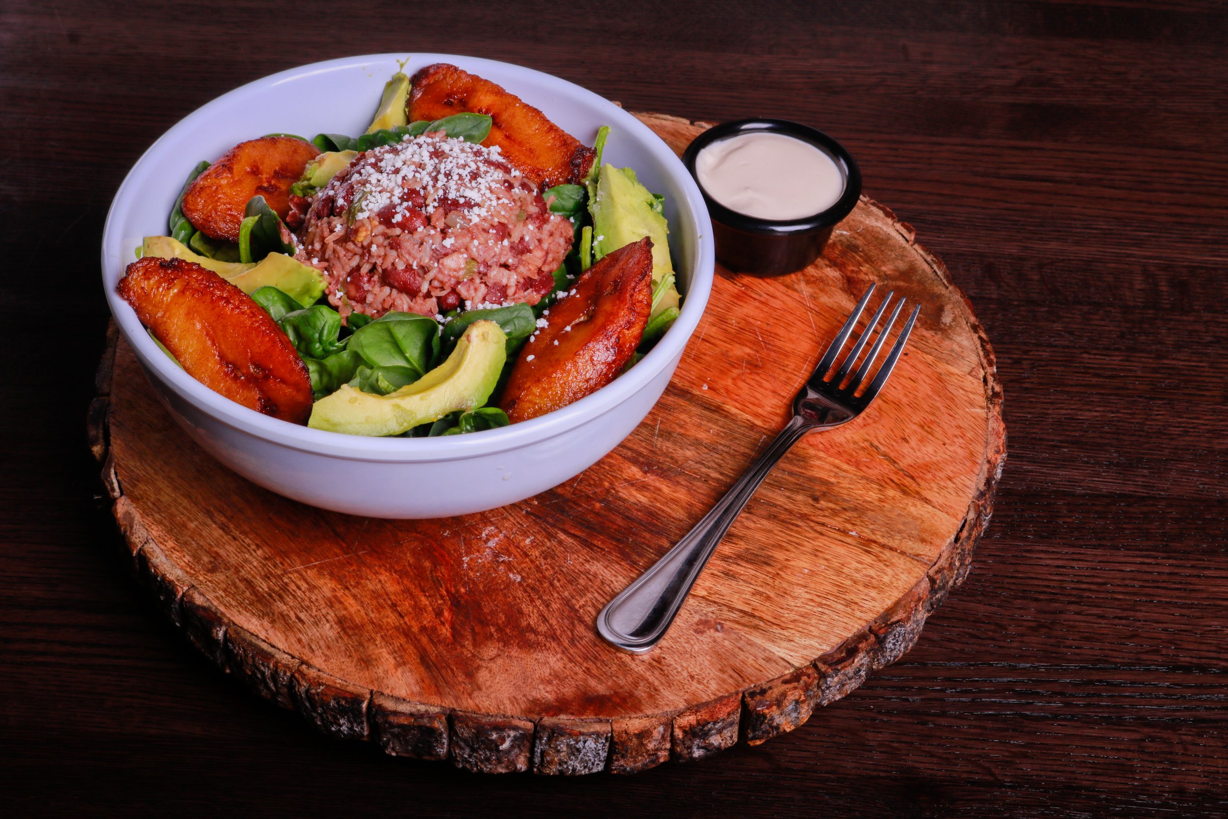 Bowl of beans and rice with avocado, leafy greens, fried plantains, and crumbled cheese, served with creamy sauce