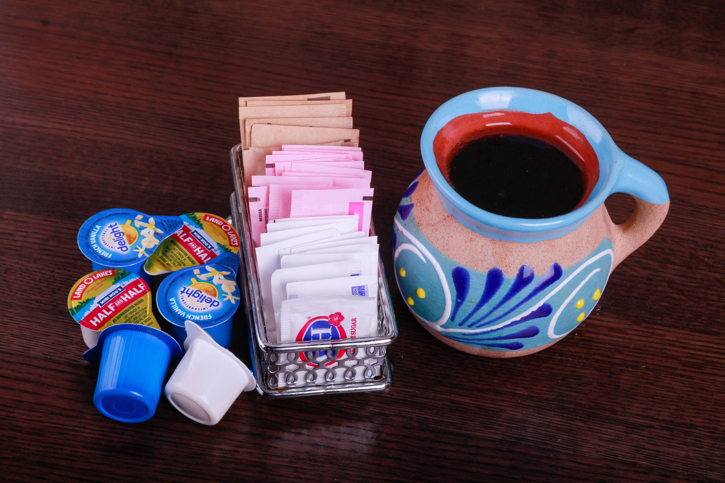 Colorful coffee mug with black coffee, sweetener packets, creamers, sugar, and plastic stirrers on a wooden table