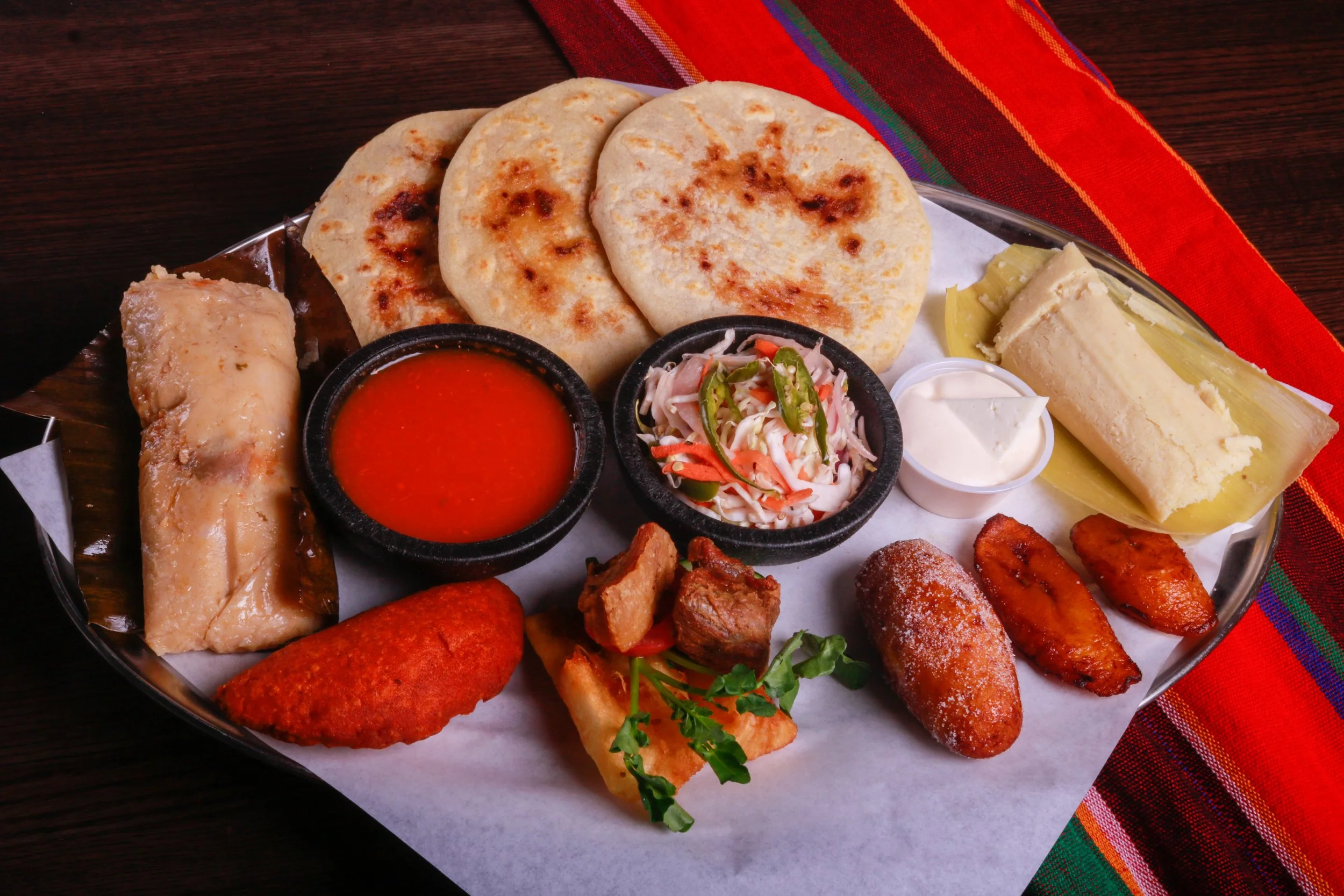 Assorted Latin American foods on a platter, including grilled tortillas, tamales, sausages, fried plantains, salad, red salsa, and crema.
