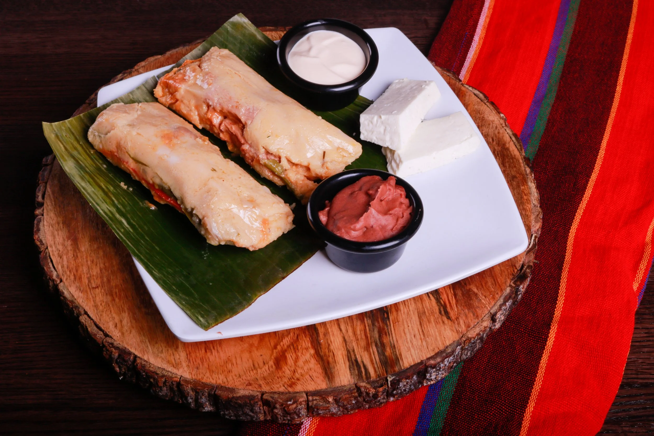 Traditional tamales served on a banana leaf with crema, queso fresco, and red salsa on a white platter