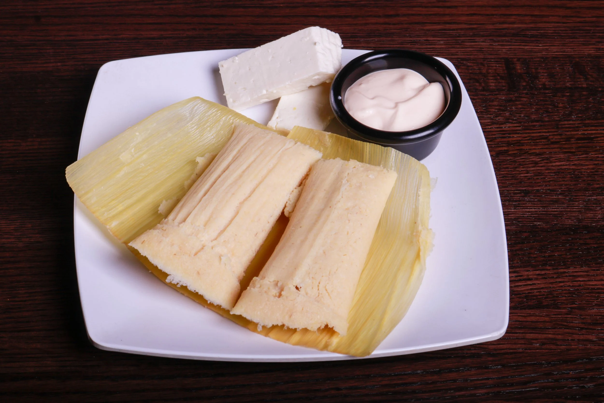 Steamed tamales served in corn husks with white cheese and sour cream on a plate