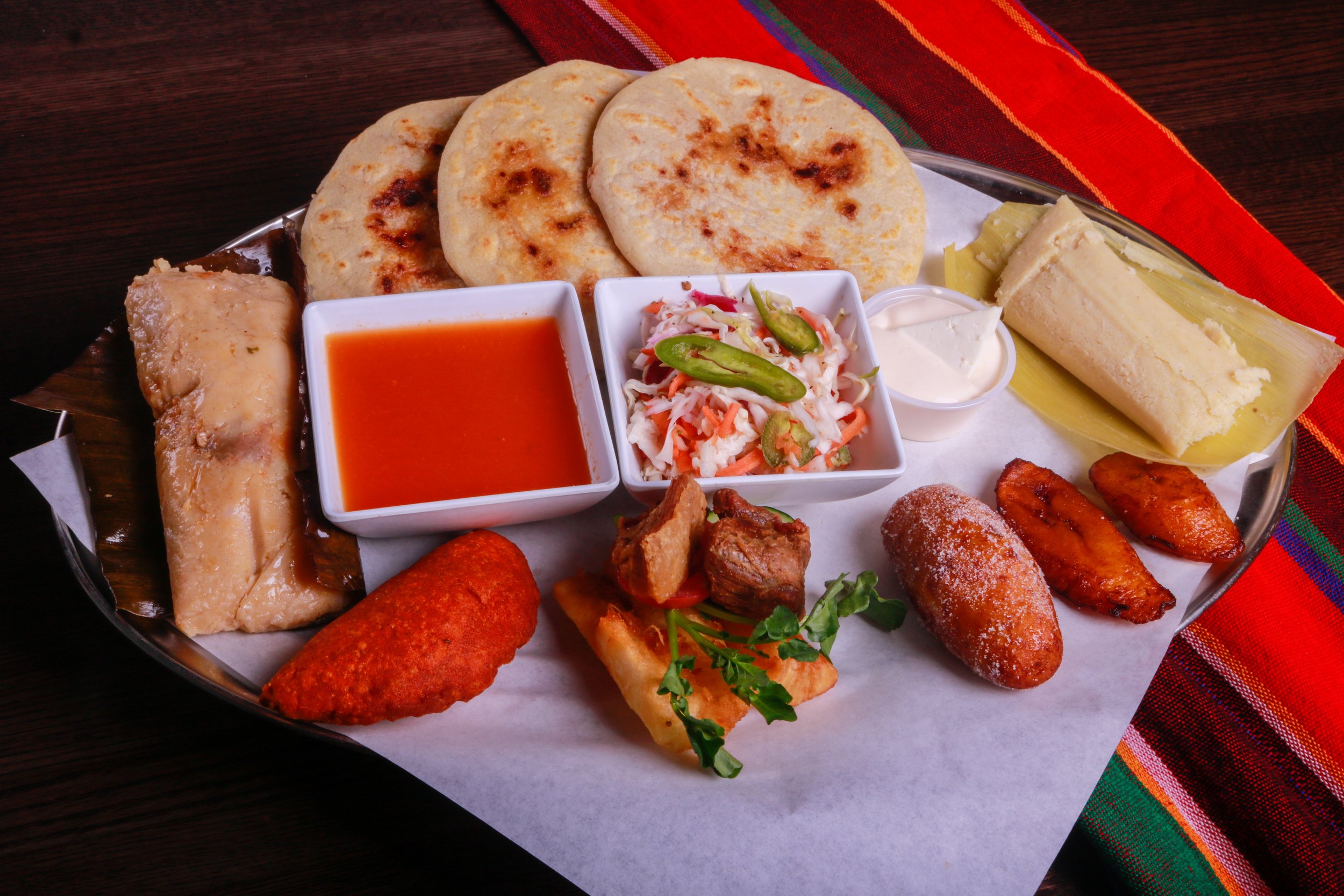 Latin American food platter with tortillas, tamales, fried plantains, meat, cabbage slaw, hot sauce, and crema