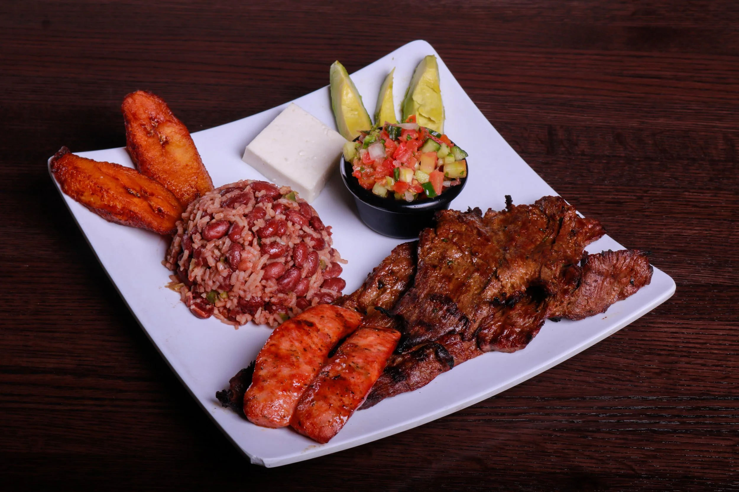 Latin American mixed grill platter with grilled meats, sausage, fried plantains, rice and beans, avocado, salsa, and white cheese