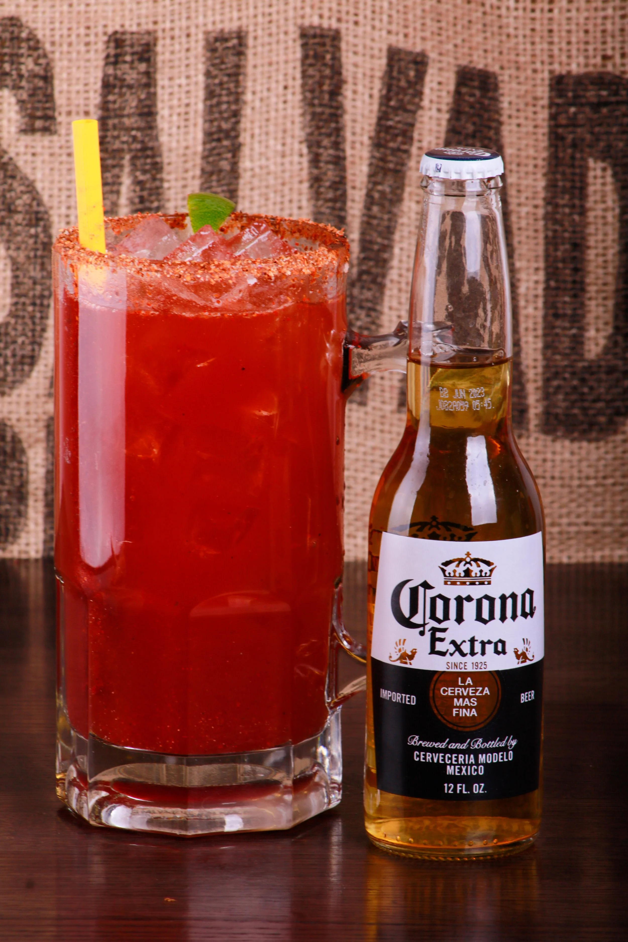 Spicy red michelada with lime, ice, chili-salt rim, and straw beside a Corona Extra beer bottle.