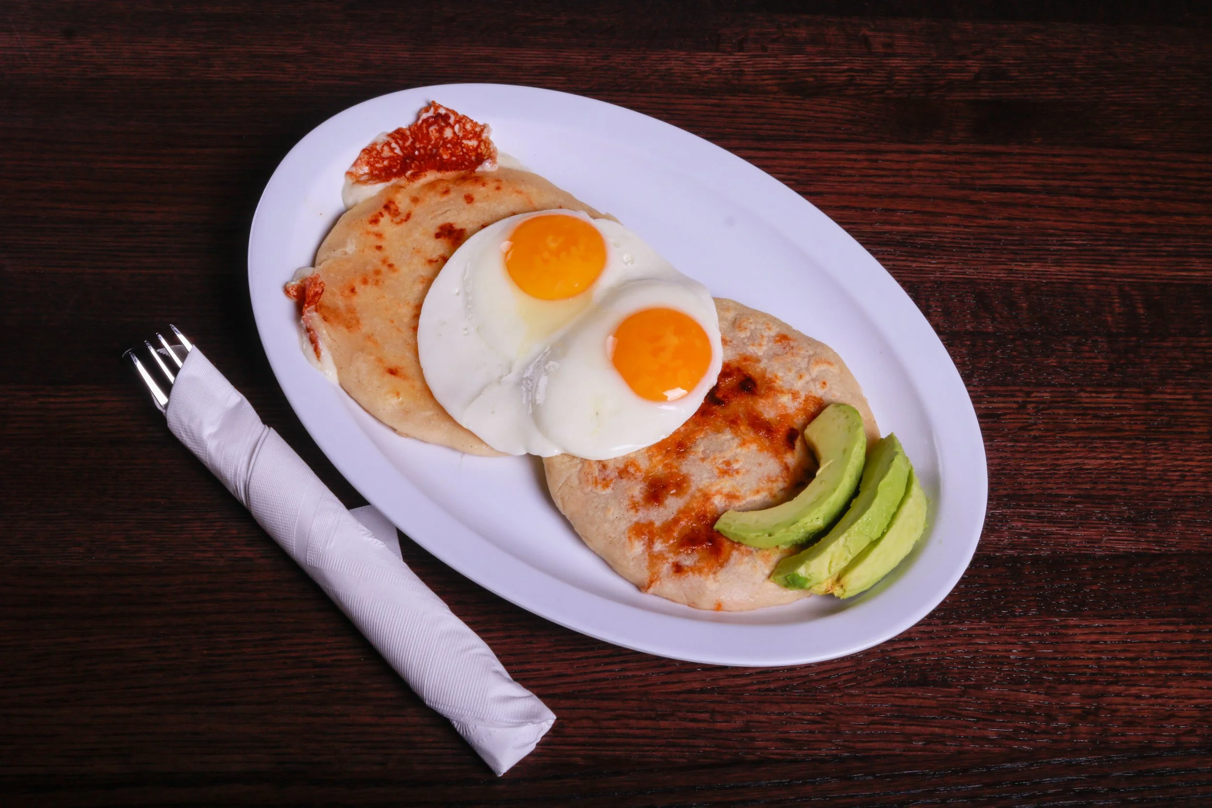 Plate of pupusas topped with two sunny-side-up eggs, red sauce, and sliced avocado