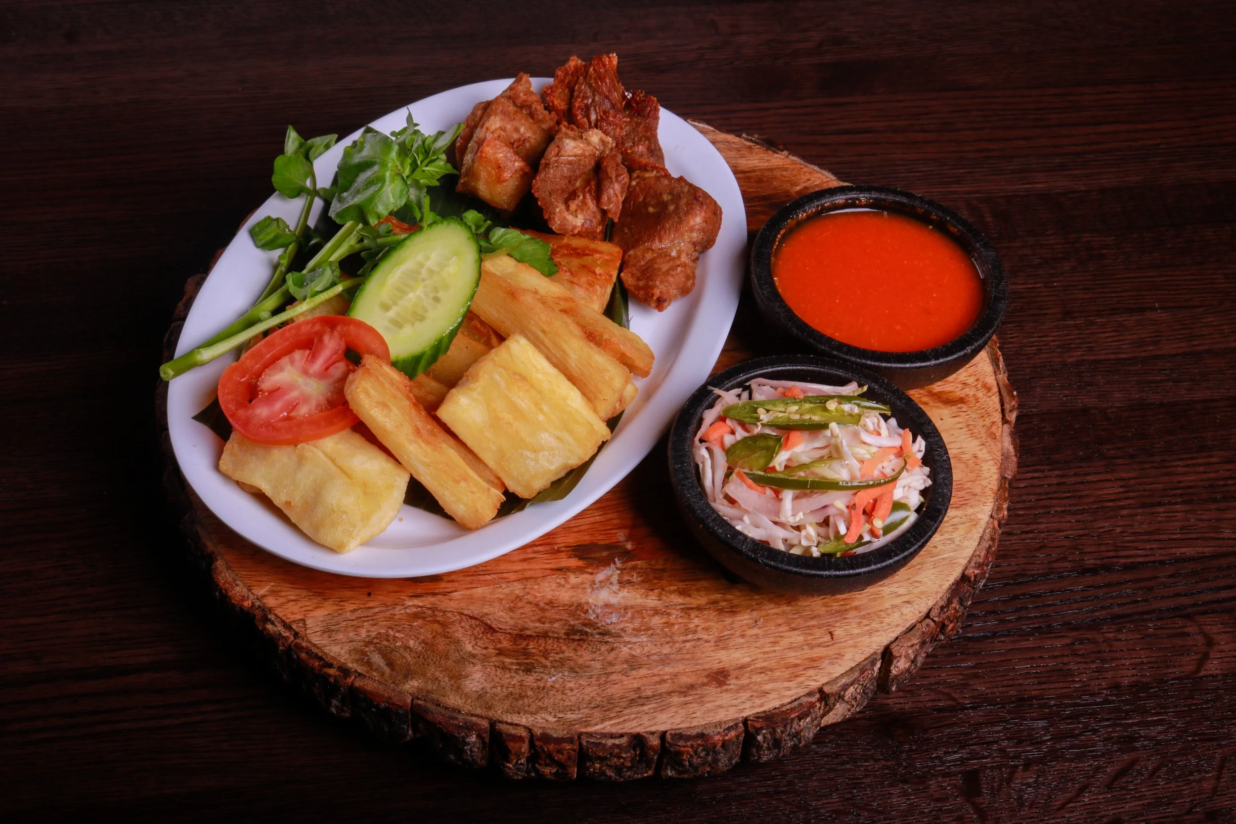 Plate of fried yuca, crispy pork, fresh vegetables, spicy red sauce, and cabbage slaw on a wooden serving board