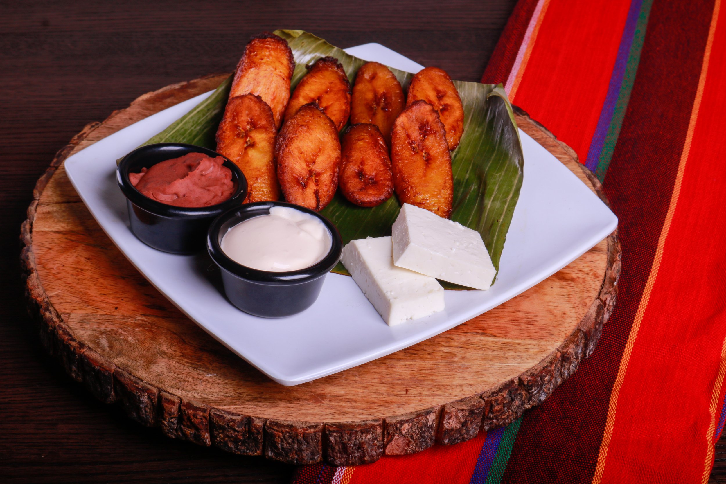 Fried ripe plantains served on a banana leaf with white cheese and two dipping sauces on a square plate